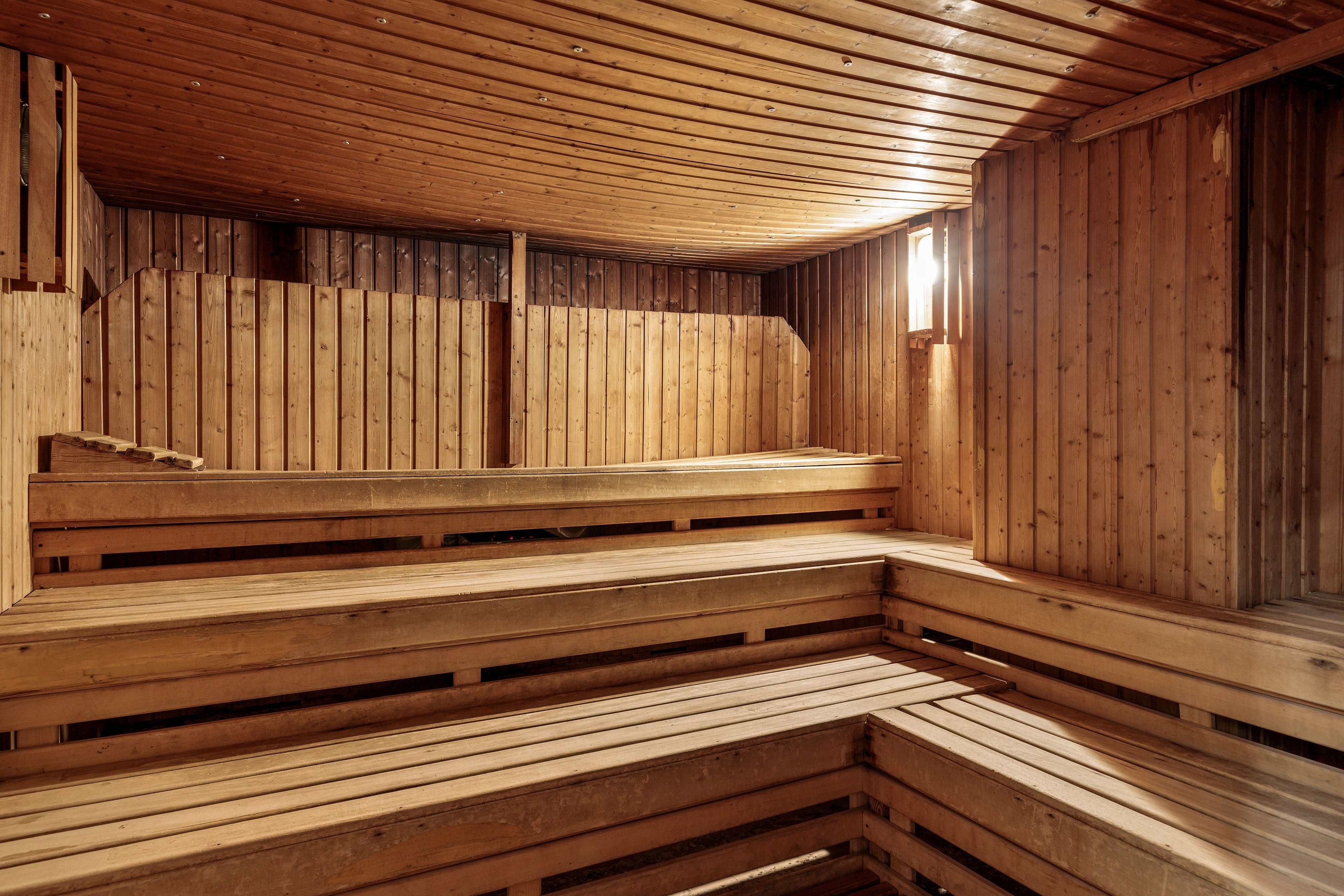 A spacious wooden sauna interior is shown with multiple tiered benches lining the walls. The room is constructed entirely of warm-toned wood planks, creating a cozy and natural atmosphere. A single wall-mounted light fixture illuminates the space, casting soft, diffused light across the benches. The sauna appears clean, empty, and ready for relaxation or spa use.