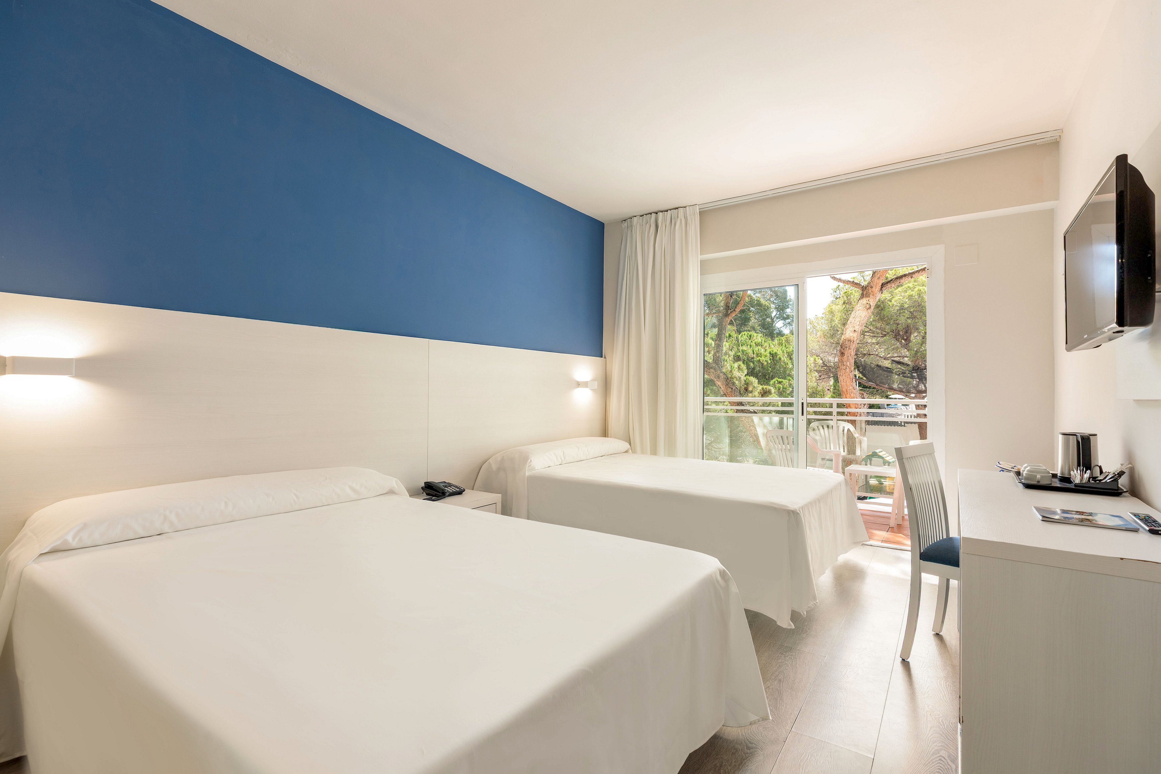 A modern hotel room features two neatly made beds with white linens against a blue accent wall. Natural light floods in through a large glass door leading to a balcony with views of green trees outside. A wall-mounted television, desk, chair, and small refreshment area complete the simple, functional furnishings. The overall atmosphere is clean, bright, and inviting, ideal for leisure or business travelers.