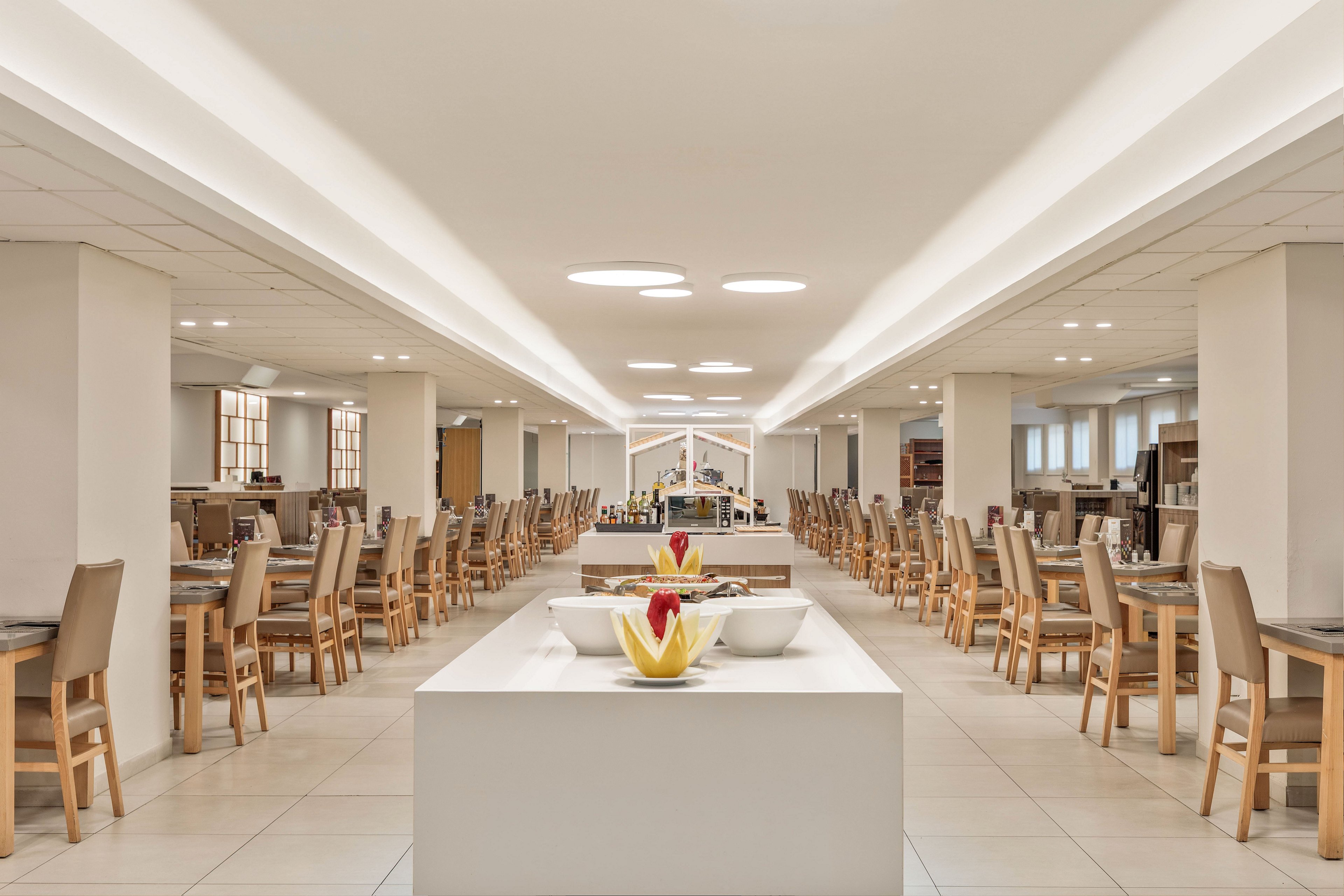 A spacious, modern cafeteria features long rows of wooden chairs and tables arranged symmetrically. In the center, a white buffet island displays bowls of fresh fruit and other dishes. Bright ceiling lights and a neutral color palette create a clean, minimalist atmosphere. The room appears ready for guests, with no people present and all seating neatly organized.