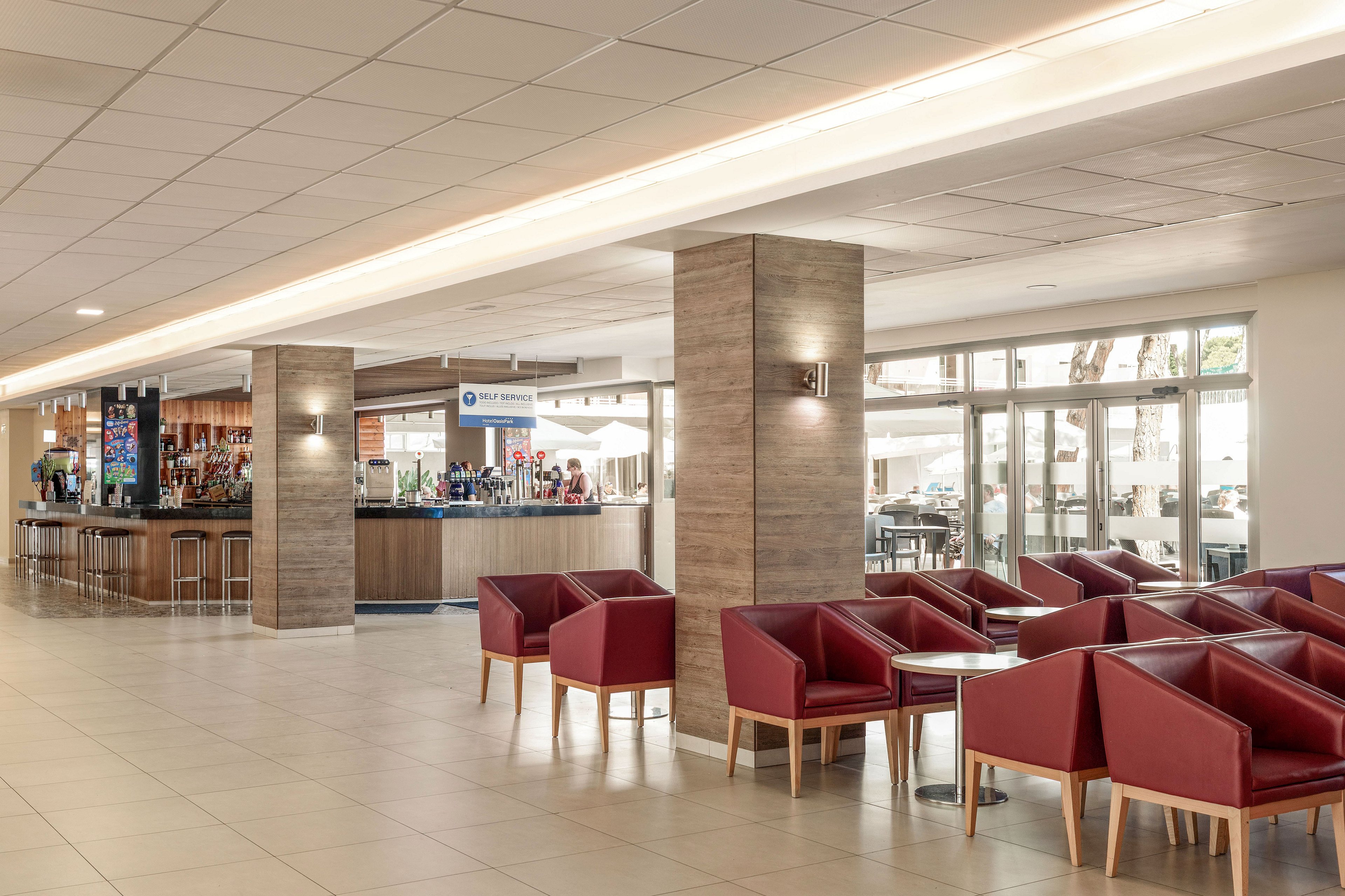 A spacious hotel lobby features a contemporary bar area with stools and a variety of bottles displayed on shelves. Red upholstered armchairs with wooden legs are arranged in seating groups on a light tiled floor. Large windows and glass doors let in natural light and offer a view of outdoor seating and trees. The overall atmosphere is clean, modern, and welcoming, suitable for socializing or relaxing.