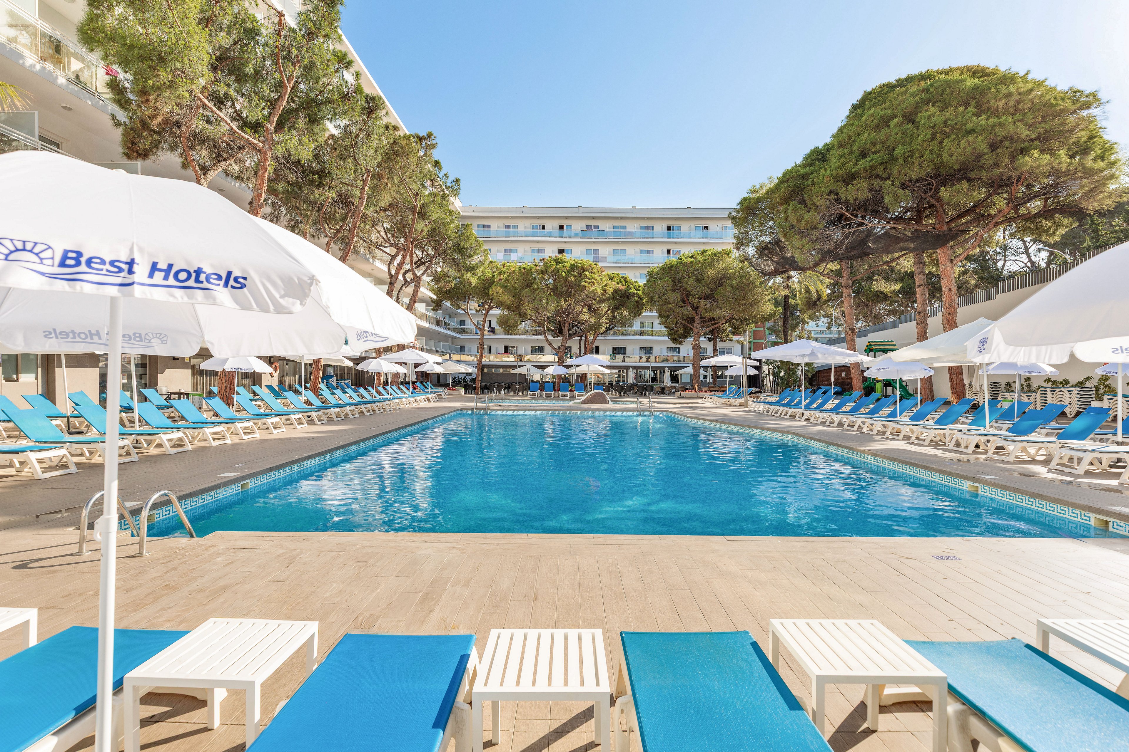 Outdoor hotel pool area with rows of blue sun loungers and white umbrellas surrounding a clear blue swimming pool. The setting appears to be a modern resort courtyard framed by tall trees and multi-story buildings. Bright daylight and clean, minimal design create a relaxed vacation atmosphere. The scene is empty of people, emphasizing tranquility and leisure.