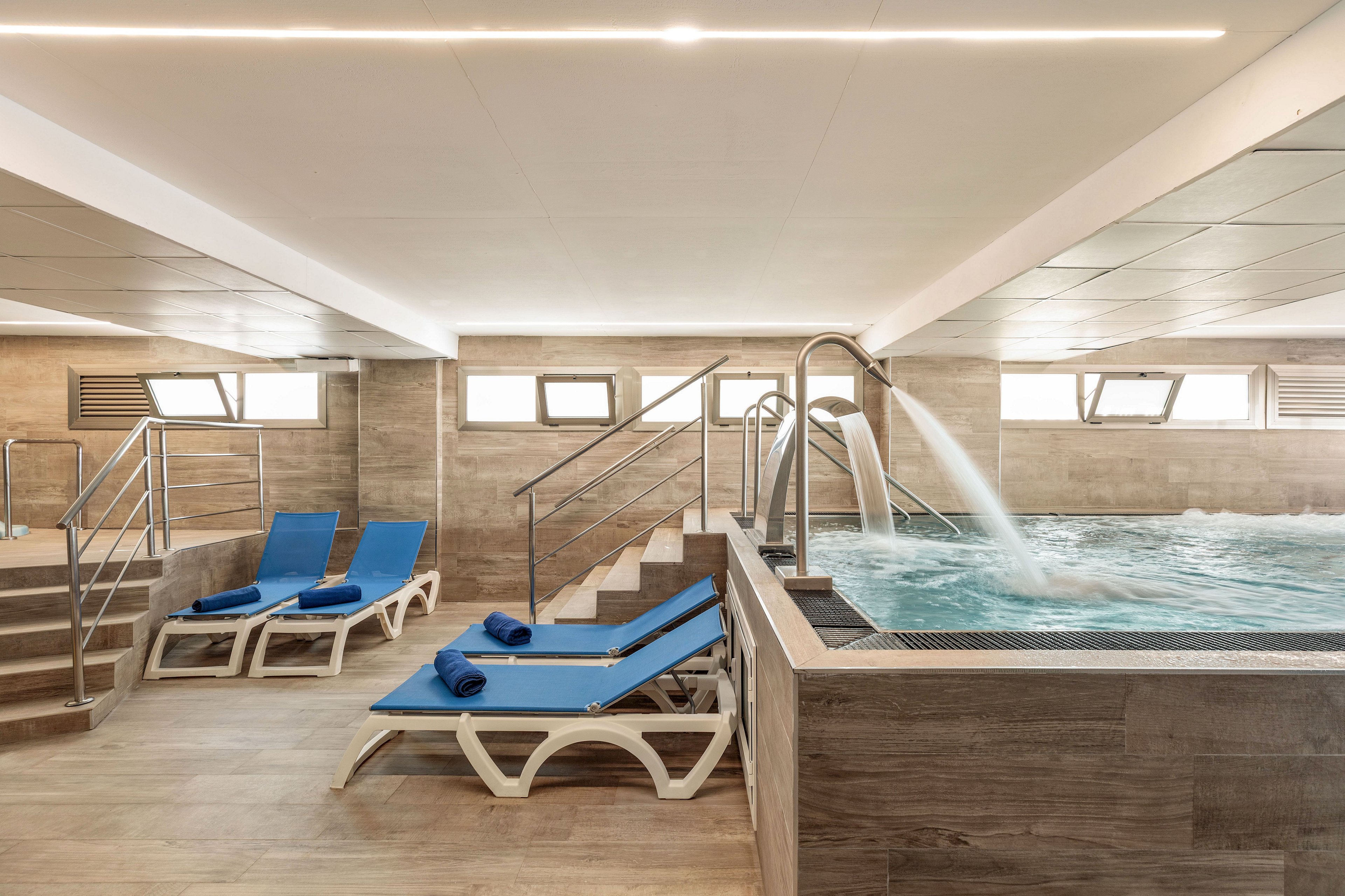 Contemporary indoor spa area featuring a rectangular hydrotherapy pool with metal handrails and water jets. Two blue cushioned loungers with rolled towels sit beside the pool on a light wood-look floor. The space is surrounded by neutral tiled walls, recessed lighting, and horizontal windows providing natural light. Overall, the scene conveys a clean, relaxing wellness environment.