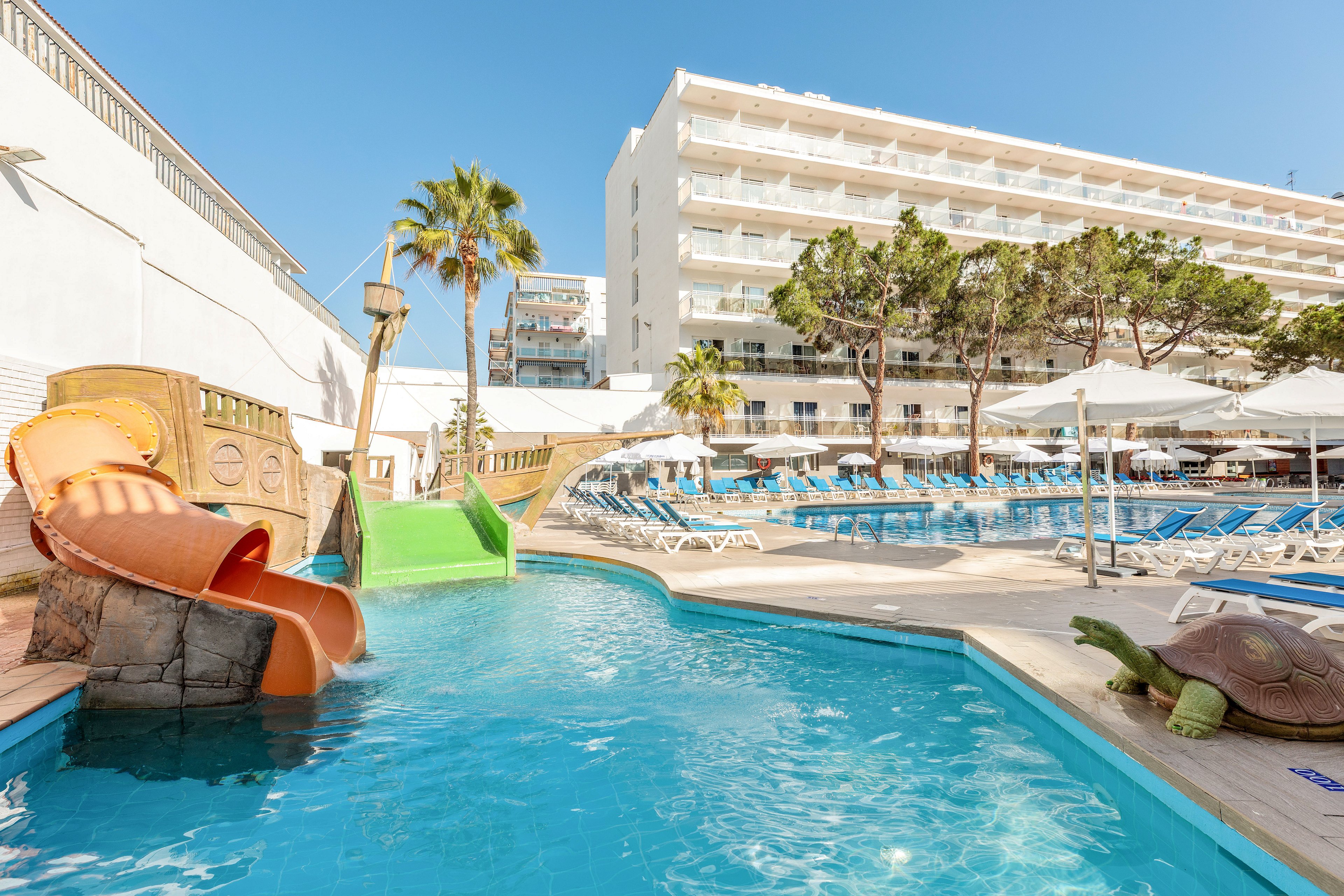 Outdoor hotel pool area featuring small curved waterslides leading into clear blue water. Sun loungers and umbrellas line the deck beside the pool, with tall palm trees and other greenery adding a tropical feel. A multi-story white hotel building rises in the background under a bright blue sky. A turtle sculpture sits near the pool edge, enhancing the playful, family-friendly atmosphere.