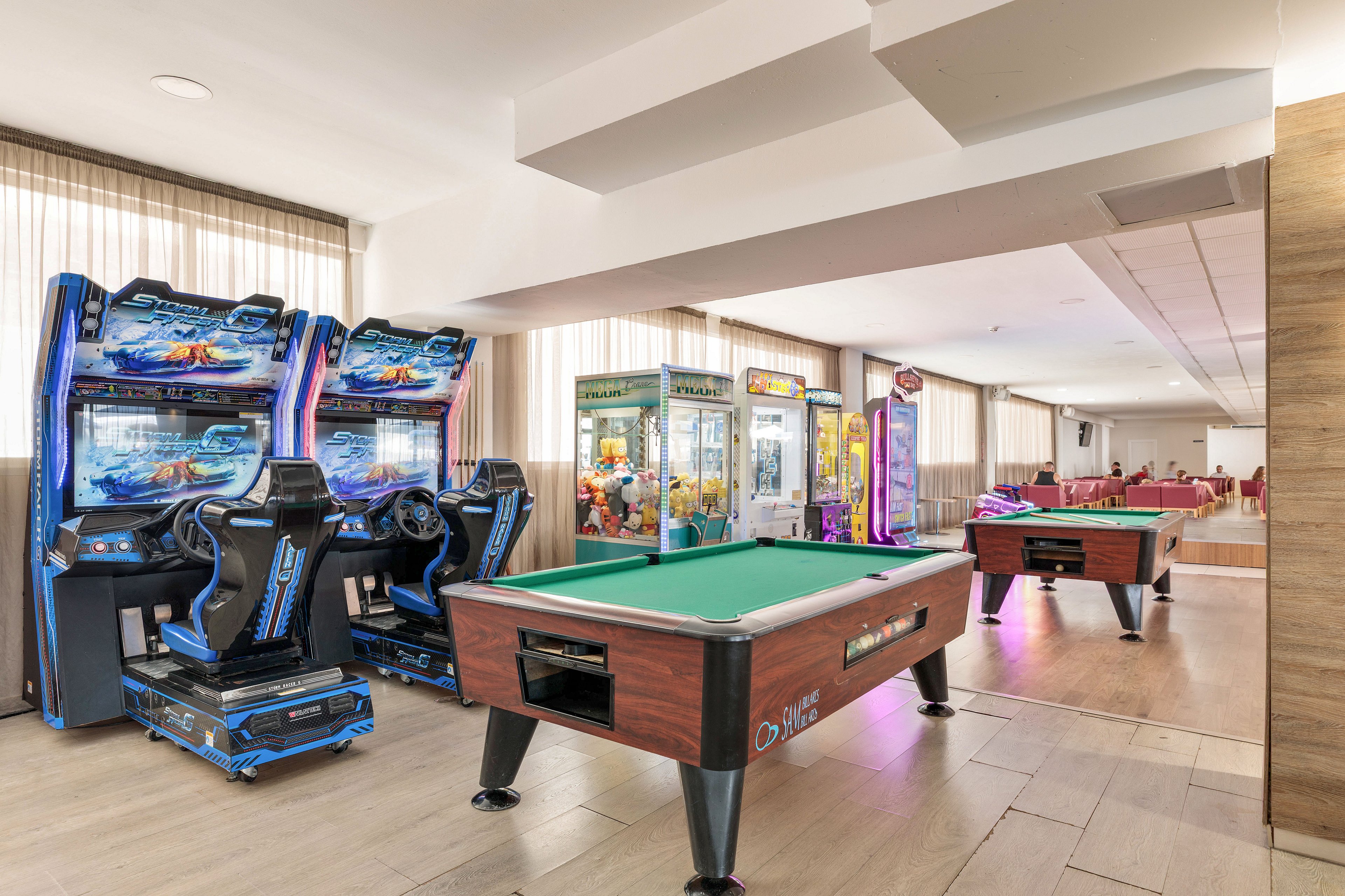 A bright indoor game room features a pool table in the foreground and several arcade machines along the wall. Claw crane games and racing simulators are visible, adding variety to the entertainment options. The space is well-lit with large windows and neutral decor, leading toward a seating area in the background. The overall atmosphere is casual and family-friendly, suitable for leisure and recreation.