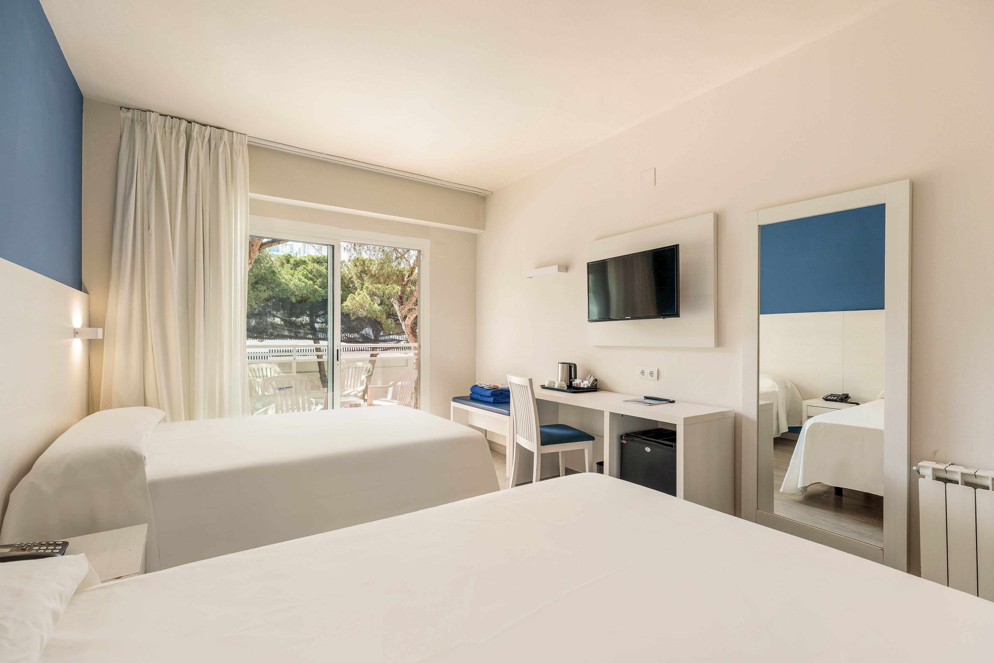 A bright, modern hotel room features two neatly made beds in a minimalist white and blue color scheme. A wall-mounted flat-screen TV faces the beds above a compact desk with a chair, electric kettle, and mini fridge. Large glass doors open to a balcony with outdoor seating and views of green trees, allowing natural light to fill the space. A full-length mirror reflects the room, enhancing the sense of openness and cleanliness.