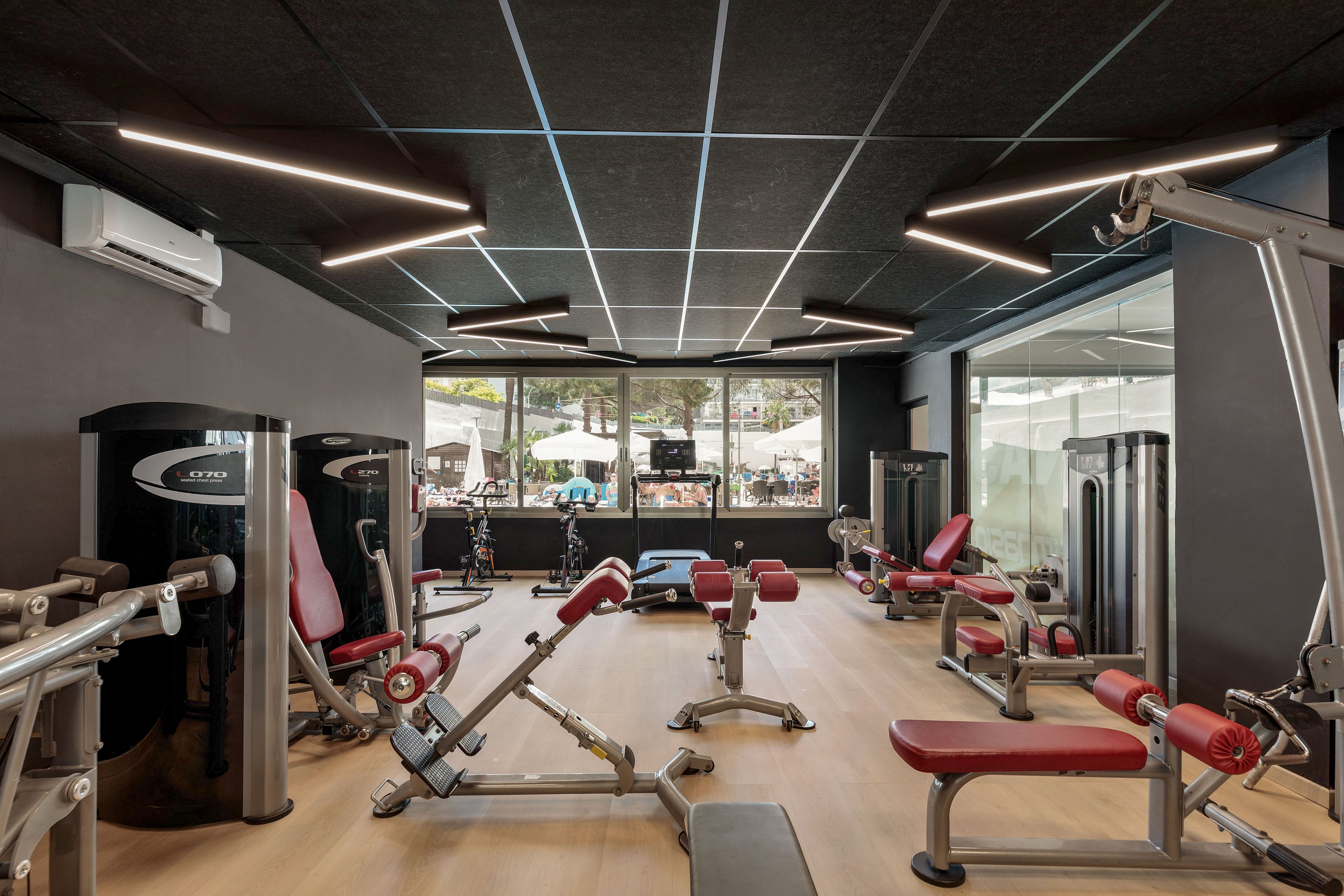 A contemporary fitness studio features multiple red and silver strength training machines arranged on a light wood floor. Large windows at the far end let in natural light and reveal an outdoor area beyond the gym. Dark walls and ceiling panels with linear LED lights create a sleek, modern atmosphere. The space appears clean, organized, and ready for individual workouts.