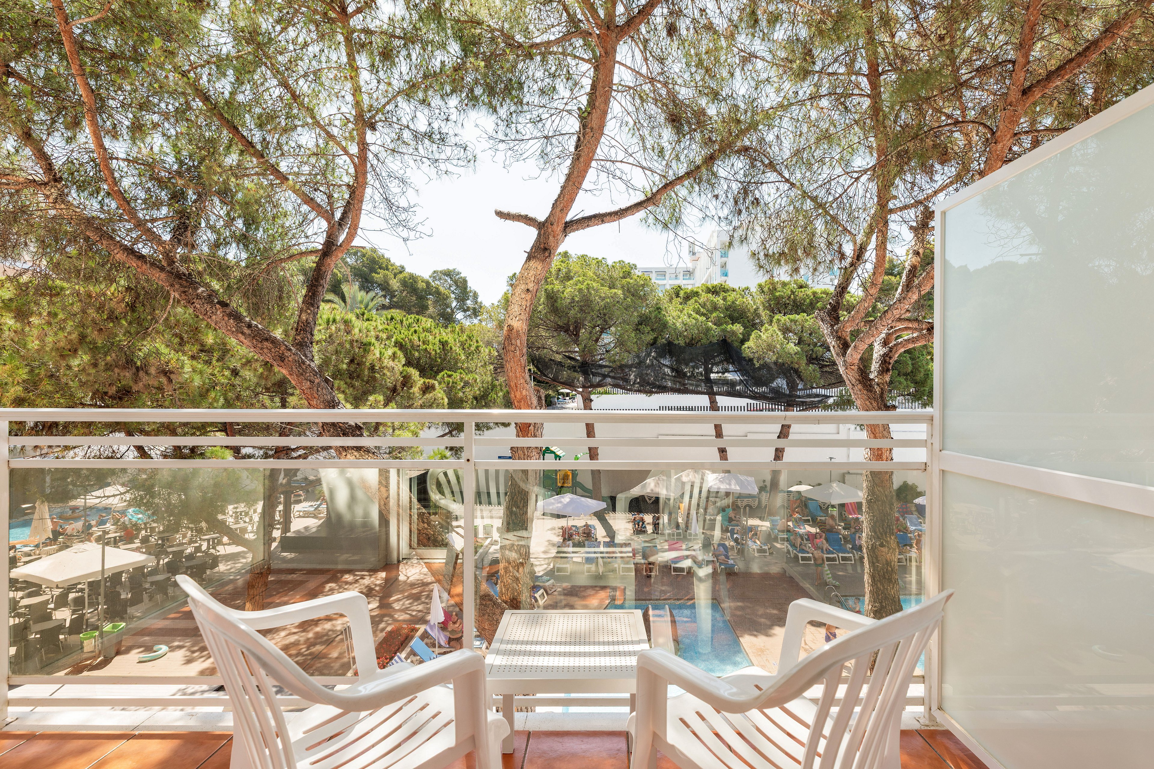 A bright balcony with two white plastic chairs overlooks a tree-filled outdoor area. Tall pine trees provide shade and frame the view beyond the glass and metal railing. Below, a poolside or patio area with sun loungers and umbrellas is partially visible. The scene suggests a relaxed holiday setting in warm, sunny weather.