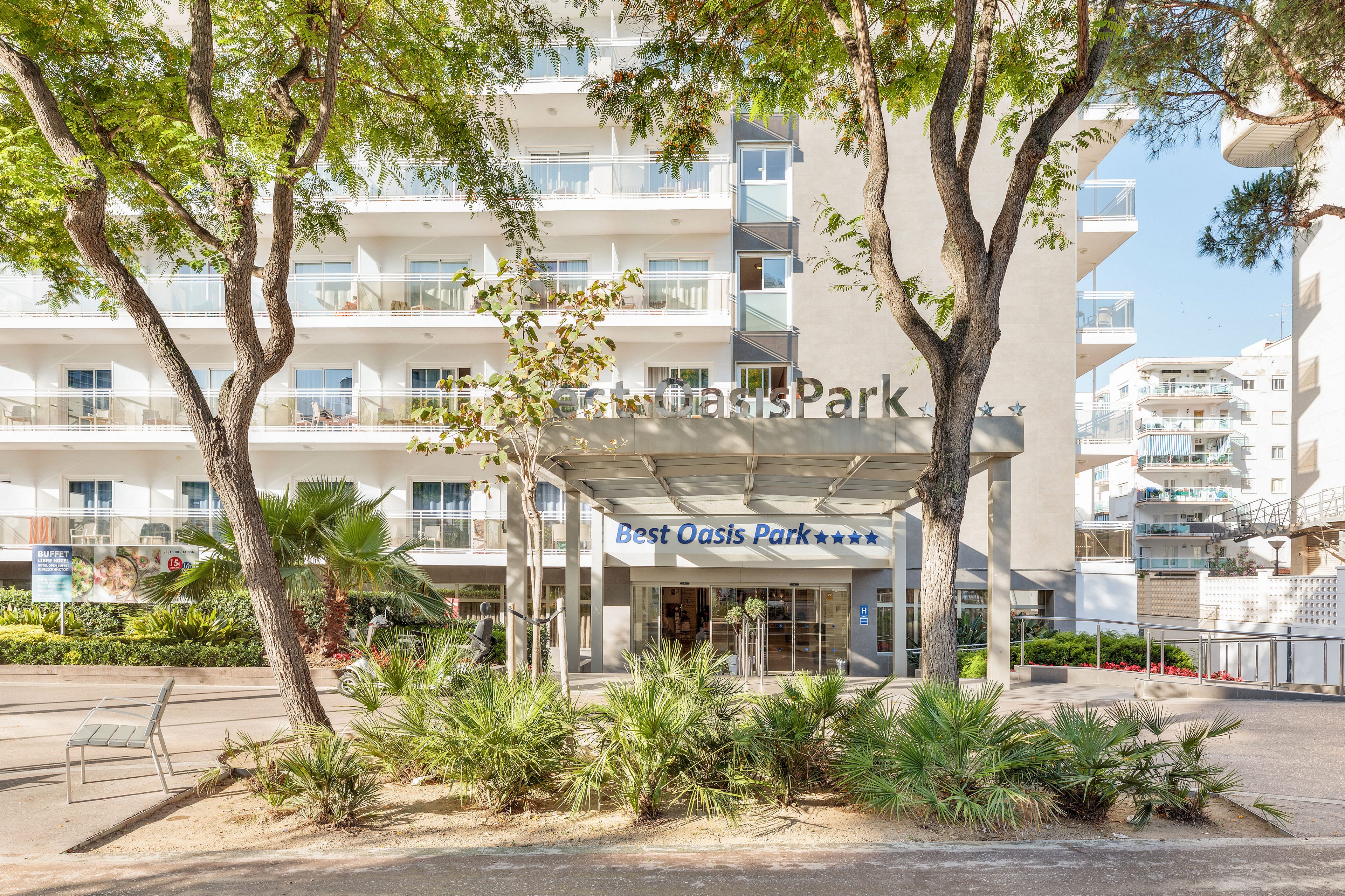 A contemporary multi-story hotel building features a glass-covered entrance labeled Best Oasis Park. The foreground includes landscaped greenery and trees framing the pathway to the main doors. Balconies with railings line the facade, suggesting guest rooms overlooking the courtyard. Bright daylight and clear skies create a welcoming, resort-like atmosphere.