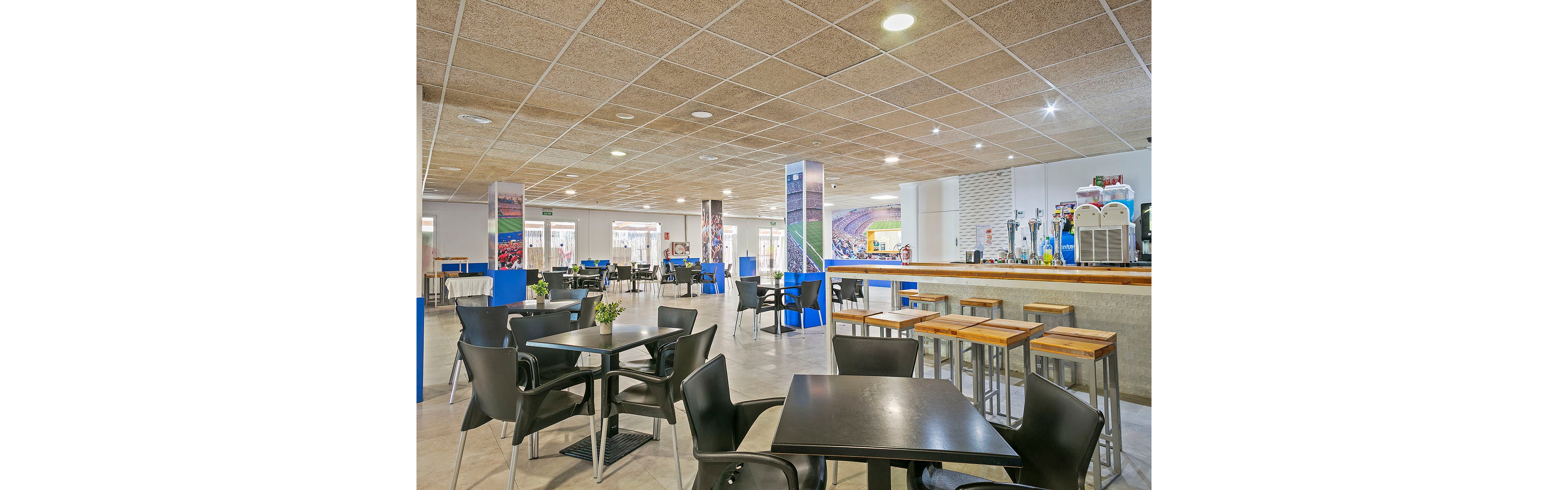 A spacious modern cafeteria features black tables and chairs arranged neatly across the room. Wooden high stools line a long bar counter equipped with beverage machines and dispensers. Bright overhead lighting and a tiled ceiling create an open, clean atmosphere. Colorful wall graphics and small plants on the tables add visual interest to the otherwise minimalist interior.