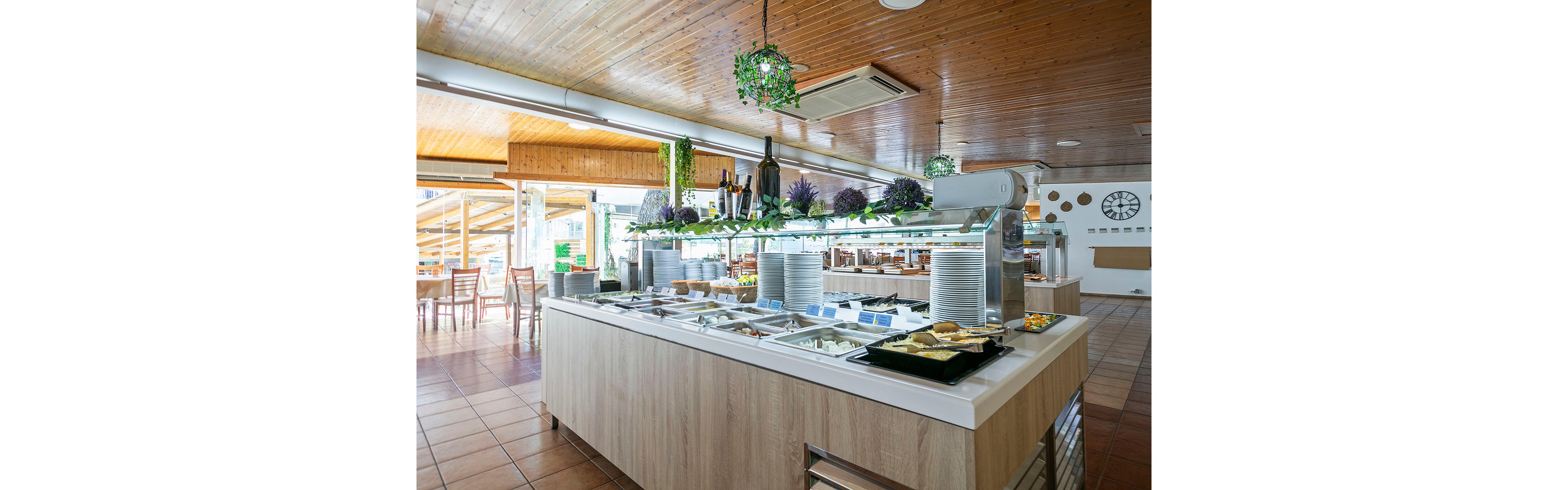 A self service buffet counter stands in the center of a bright restaurant dining area. Stainless steel food trays, plates, and utensils are neatly arranged under a glass sneeze guard. Decorative greenery, bottles, and purple flowers line the top of the counter, complementing the warm wooden ceiling and tiled floor. Large windows and open seating in the background create an airy, casual dining atmosphere.