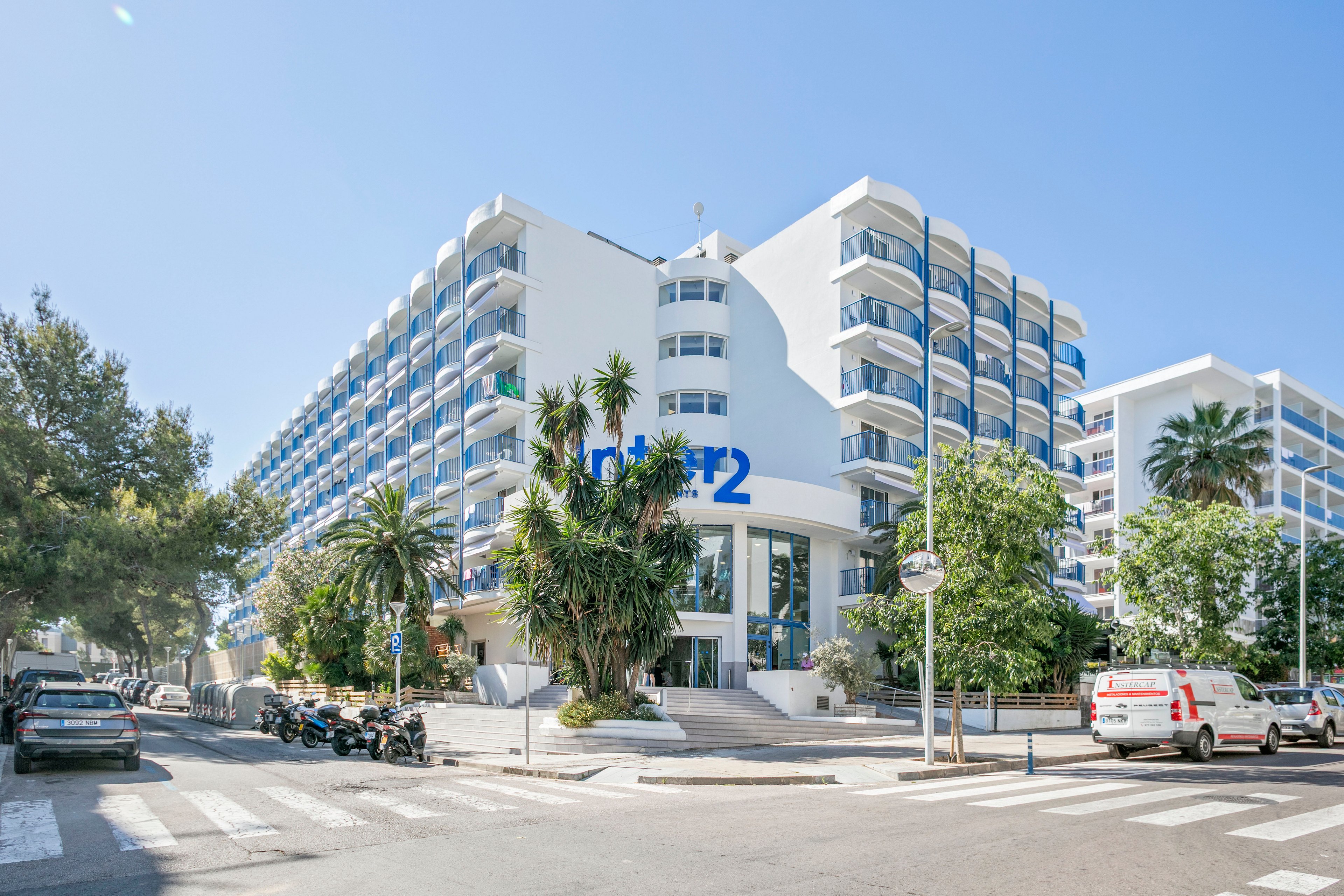 A large modern hotel building with multiple floors and rows of balconies stands on a sunny street corner. The white facade and blue balcony railings give the structure a bright, coastal appearance. Palm trees and other greenery frame the entrance, while parked cars and scooters line the surrounding streets. Clear blue skies and strong daylight suggest a warm, pleasant climate ideal for vacation stays.