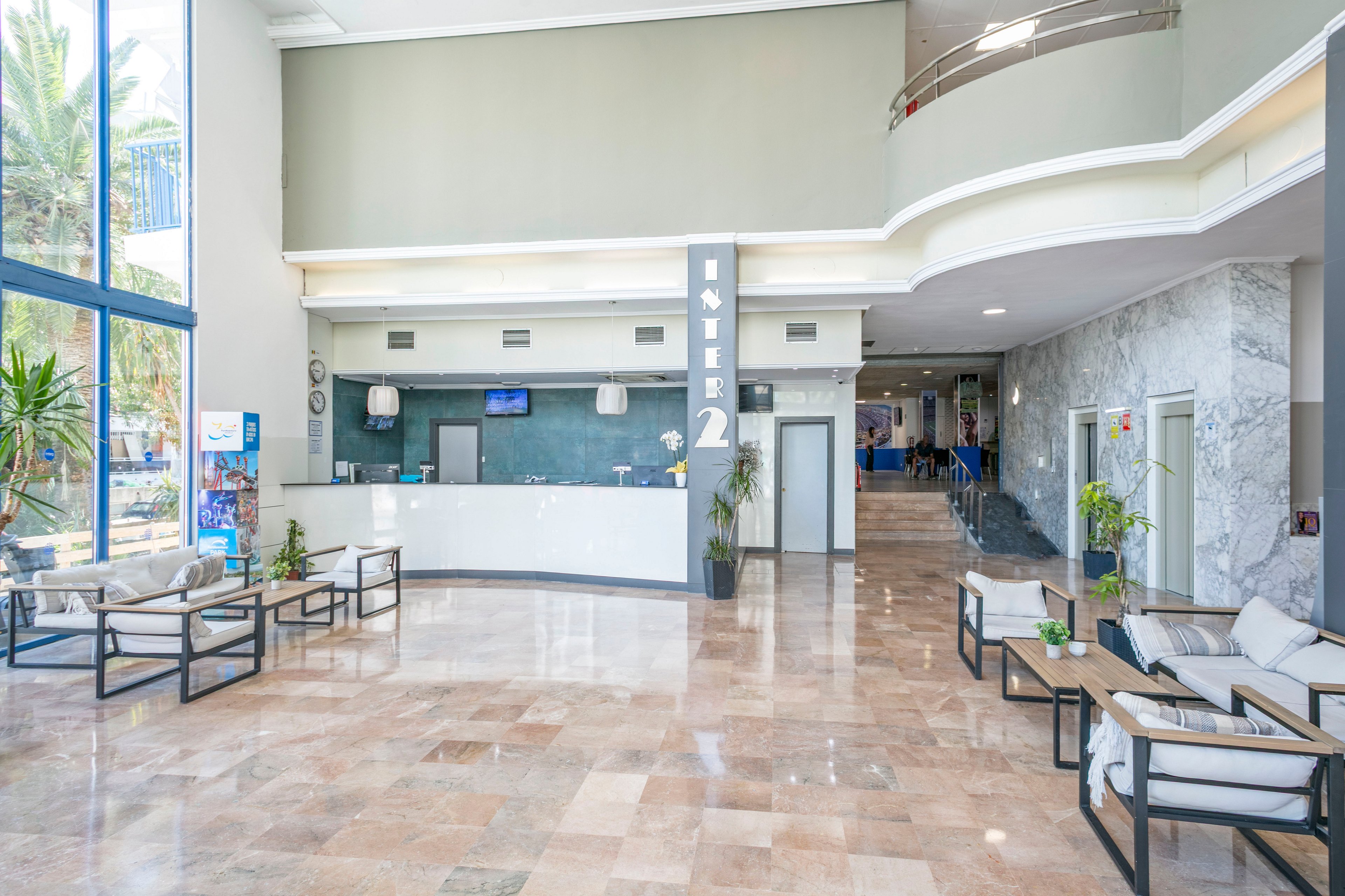 A spacious hotel lobby features a modern reception desk with a clean, minimalist design. Light marble floors and neutral-colored walls create an airy, welcoming atmosphere. Comfortable seating areas with cushioned chairs and small tables are arranged near large windows that let in natural light. Green plants and subtle decorative elements add warmth to the contemporary interior.