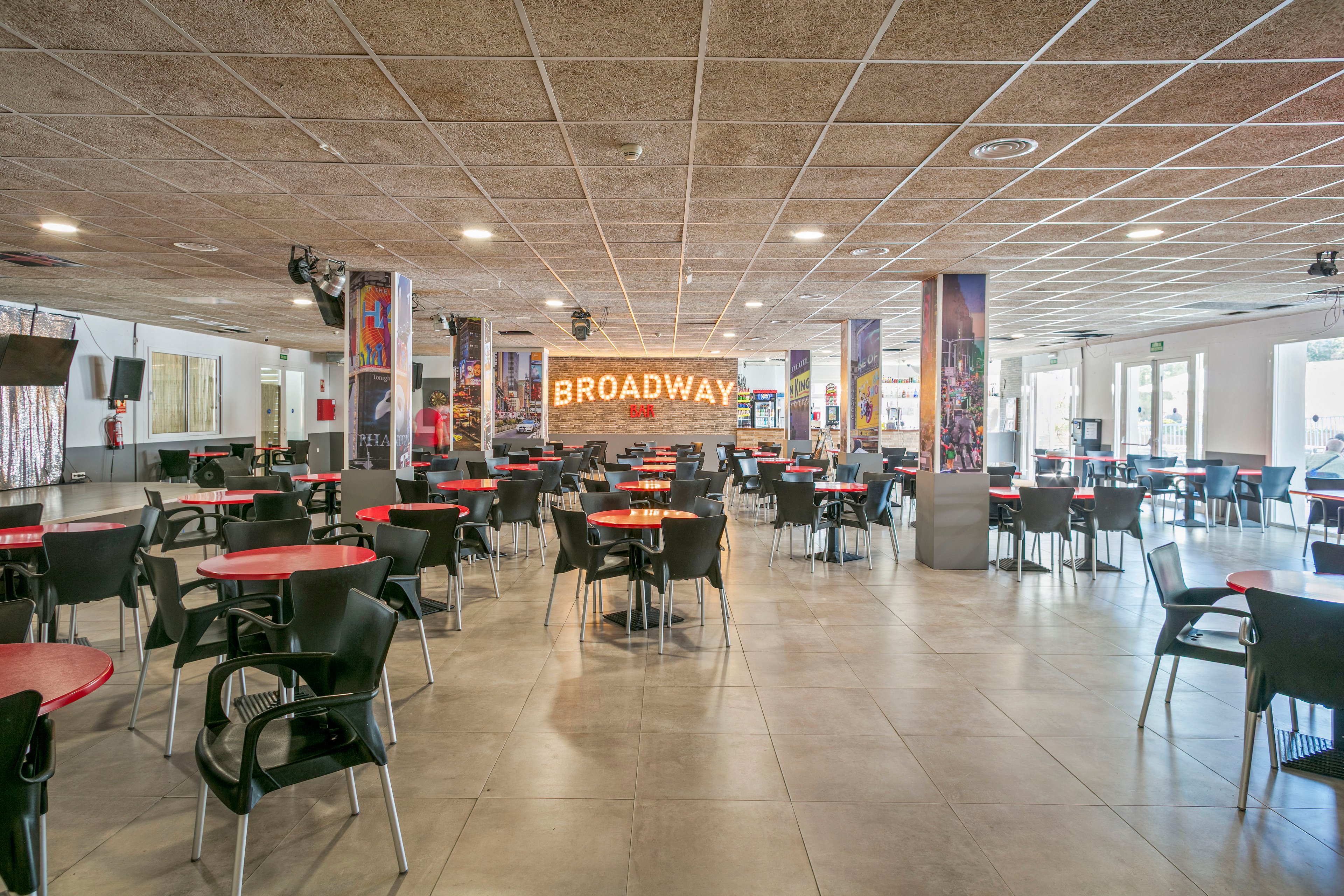 A large, modern cafeteria is filled with rows of black chairs and round tables with red tops. The space is brightly lit with a grid of ceiling lights and has a neutral tiled floor. A prominent illuminated sign reading "BROADWAY" is mounted on the back wall, flanked by tall decorative columns. Large windows and glass doors on the right side allow natural light into the open, empty dining area.