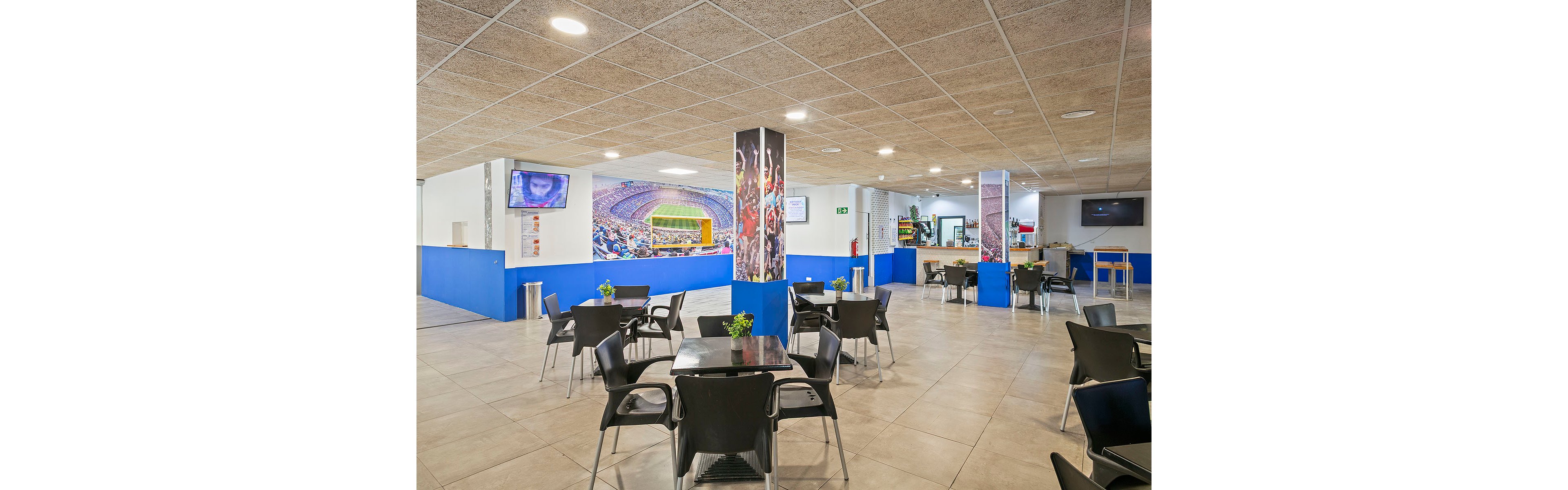 A spacious cafeteria area features multiple black tables and chairs arranged on a tiled floor. Blue counters and wall panels frame the space, with a food or drink service counter visible in the background. Large sports-themed murals and televisions showing a stadium scene emphasize the athletic atmosphere. Bright lighting and clean, open layout suggest a casual, family-friendly environment suitable for fans before or after a game.
