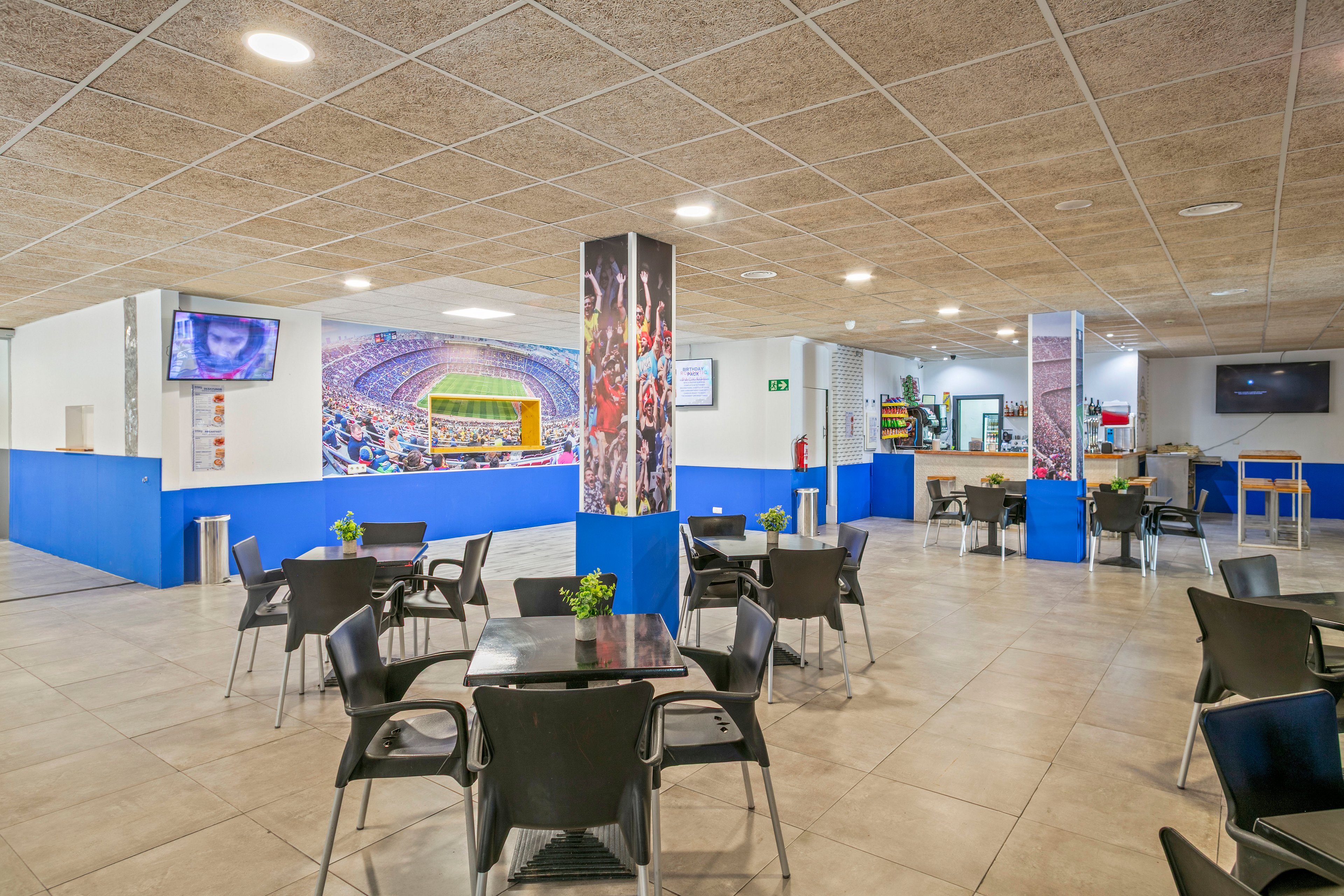 Spacious cafeteria seating is arranged in a modern sports stadium interior. Black chairs and glass-topped tables are spread across a tiled floor with bright blue wall accents. Large columns feature colorful images of cheering crowds and a lit stadium field. Multiple wall-mounted screens and a service counter are visible in the background, creating a casual game-day atmosphere.