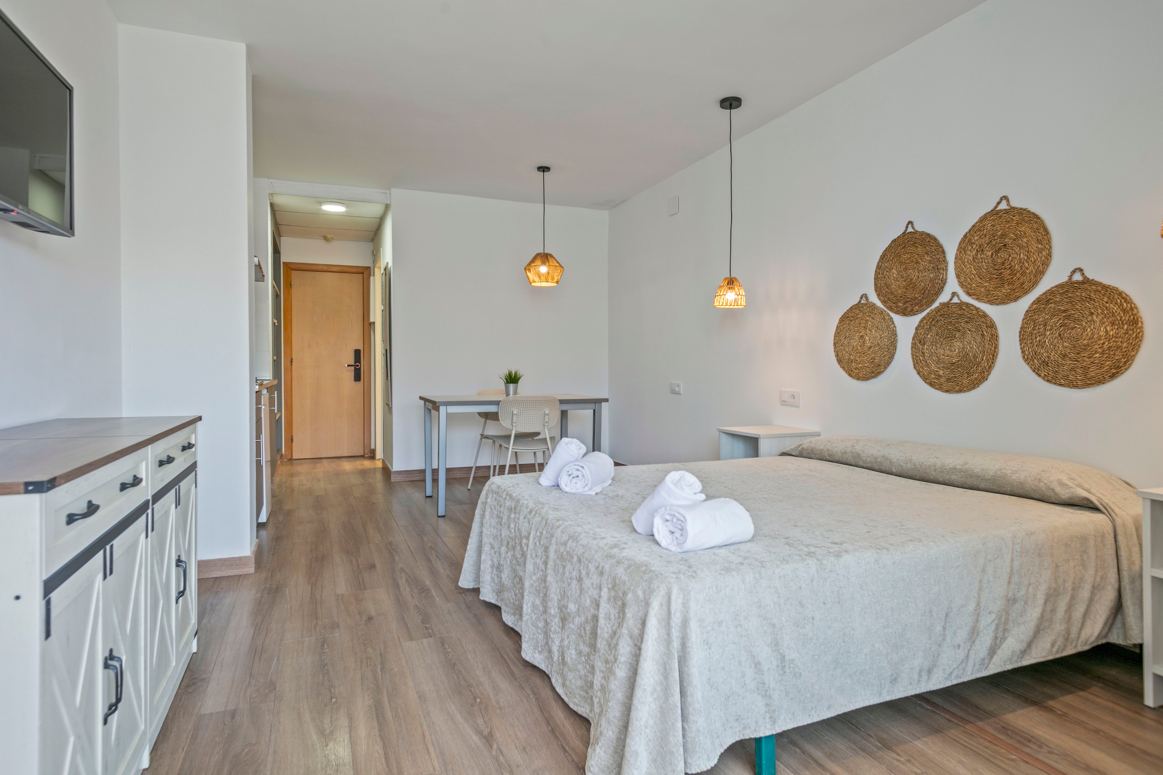 A bright, minimalist hotel room features a neatly made double bed with folded white towels placed on top. The space includes light wood flooring, a wall-mounted television, and a sideboard along one wall. In the background, a small dining or work table with chairs sits near the entrance door, illuminated by two hanging pendant lights. Woven wall decor and neutral bedding create a calm, contemporary atmosphere suitable for guests.