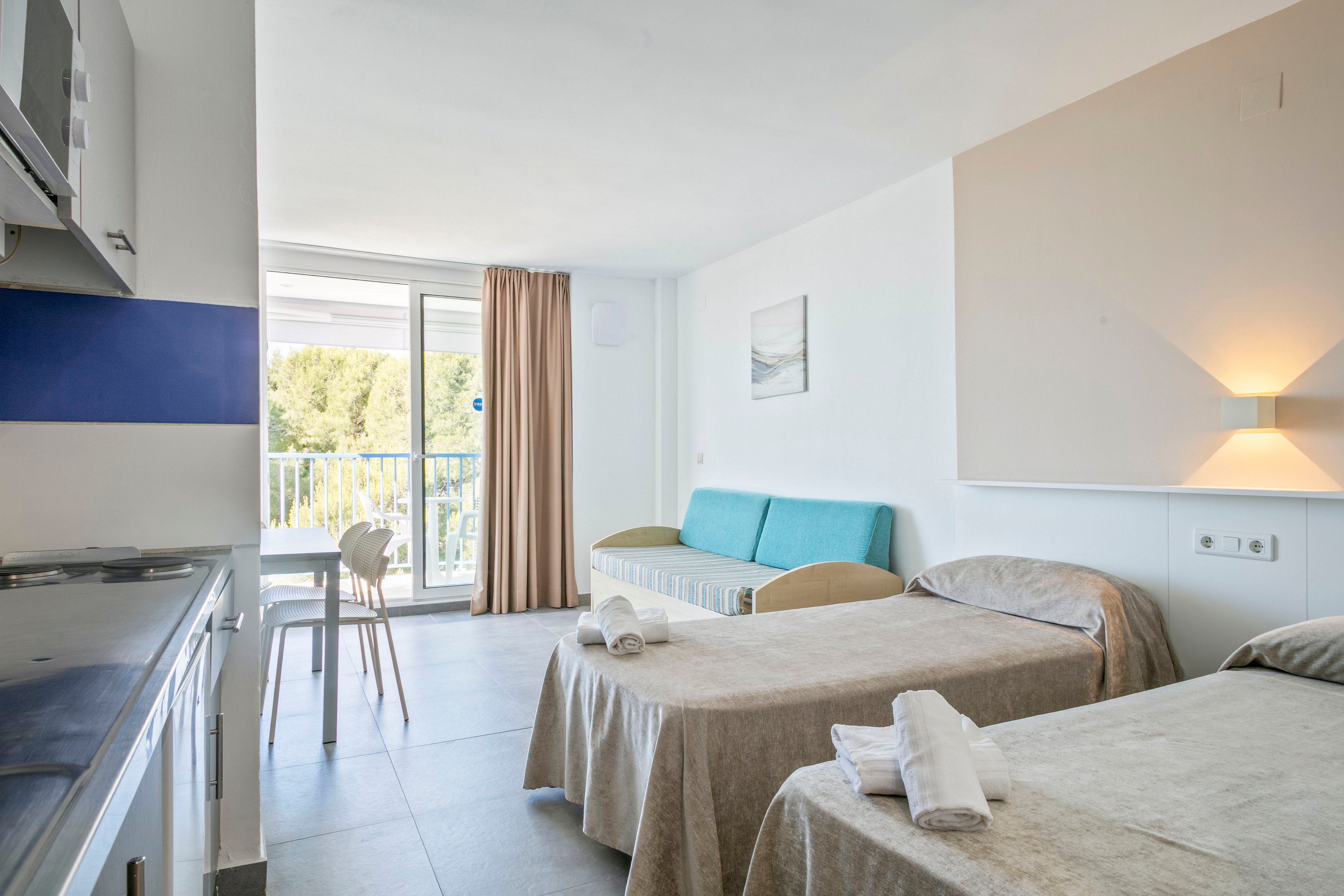 A bright, modern hotel room features two single beds neatly made with beige linens and folded white towels. To the side, a compact kitchenette and dining table face a large sliding glass door that opens to a balcony. A turquoise sofa and simple wall art add subtle color to the otherwise neutral decor. Natural light floods the space, creating an airy, minimalist atmosphere with views of greenery outside.