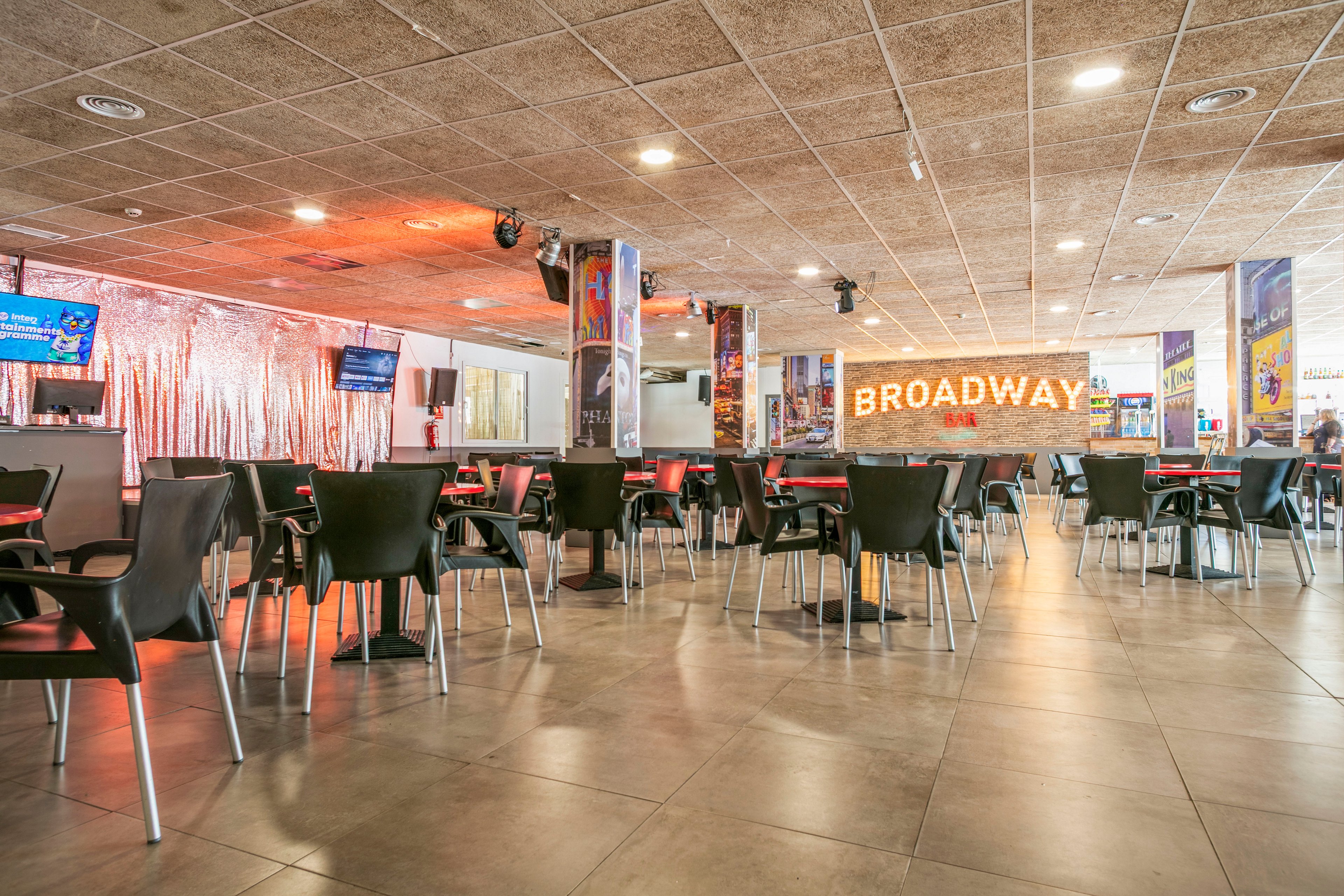 A spacious modern cafe features rows of black chairs and small round tables arranged across a tiled floor. Warm lighting and a reflective metallic curtain create a cozy yet contemporary atmosphere. A large illuminated sign reading "BROADWAY" dominates the background wall, adding a theatrical theme to the space. Digital screens and colorful posters enhance the lively, entertainment-inspired setting.