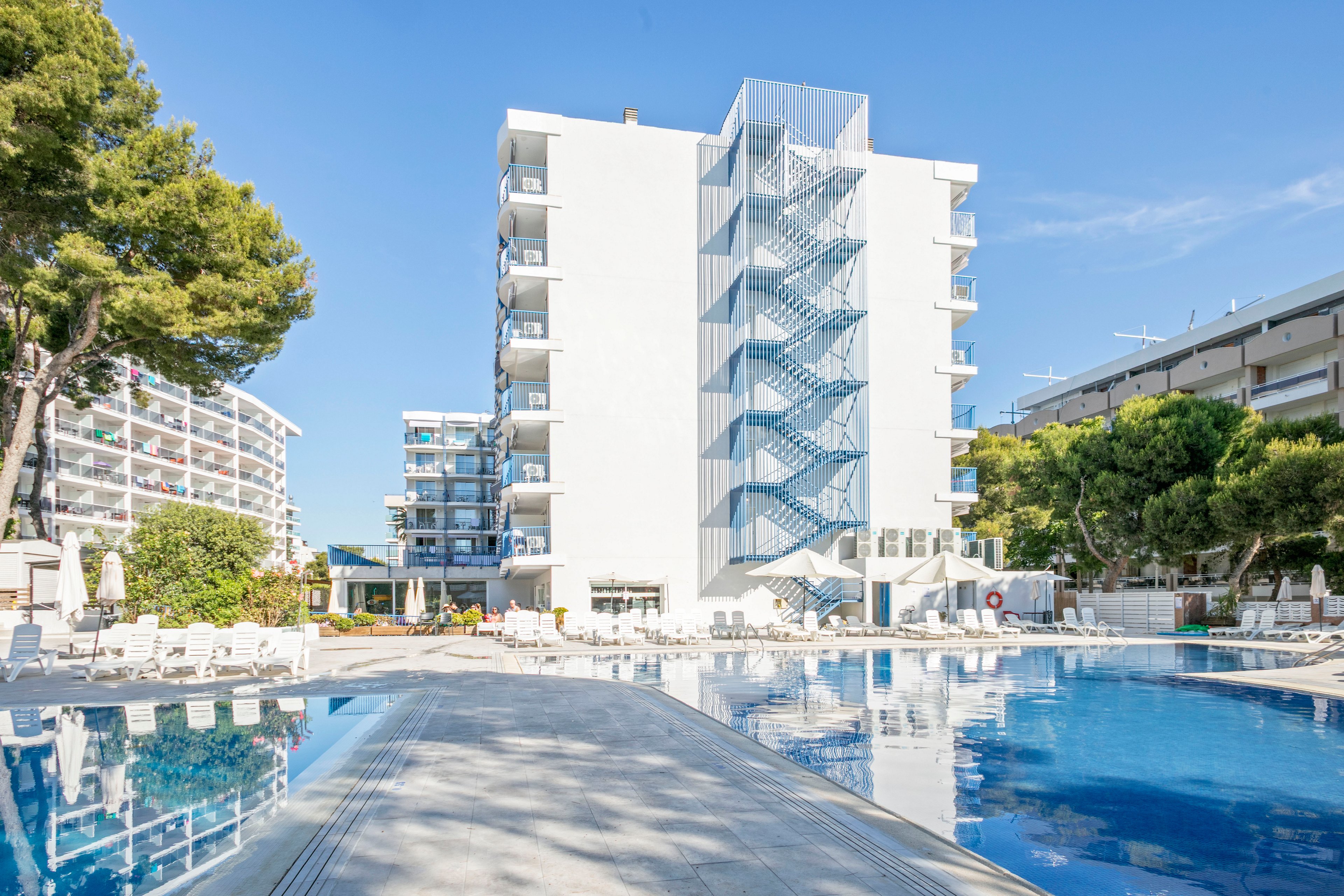 A modern white hotel building stands beside a large outdoor swimming pool under a clear blue sky. Sun loungers and umbrellas are arranged around the pool, suggesting a relaxing vacation setting. Surrounding trees and neighboring buildings frame the scene, adding greenery and urban resort context. The bright daylight and reflections in the water create a clean, airy atmosphere.