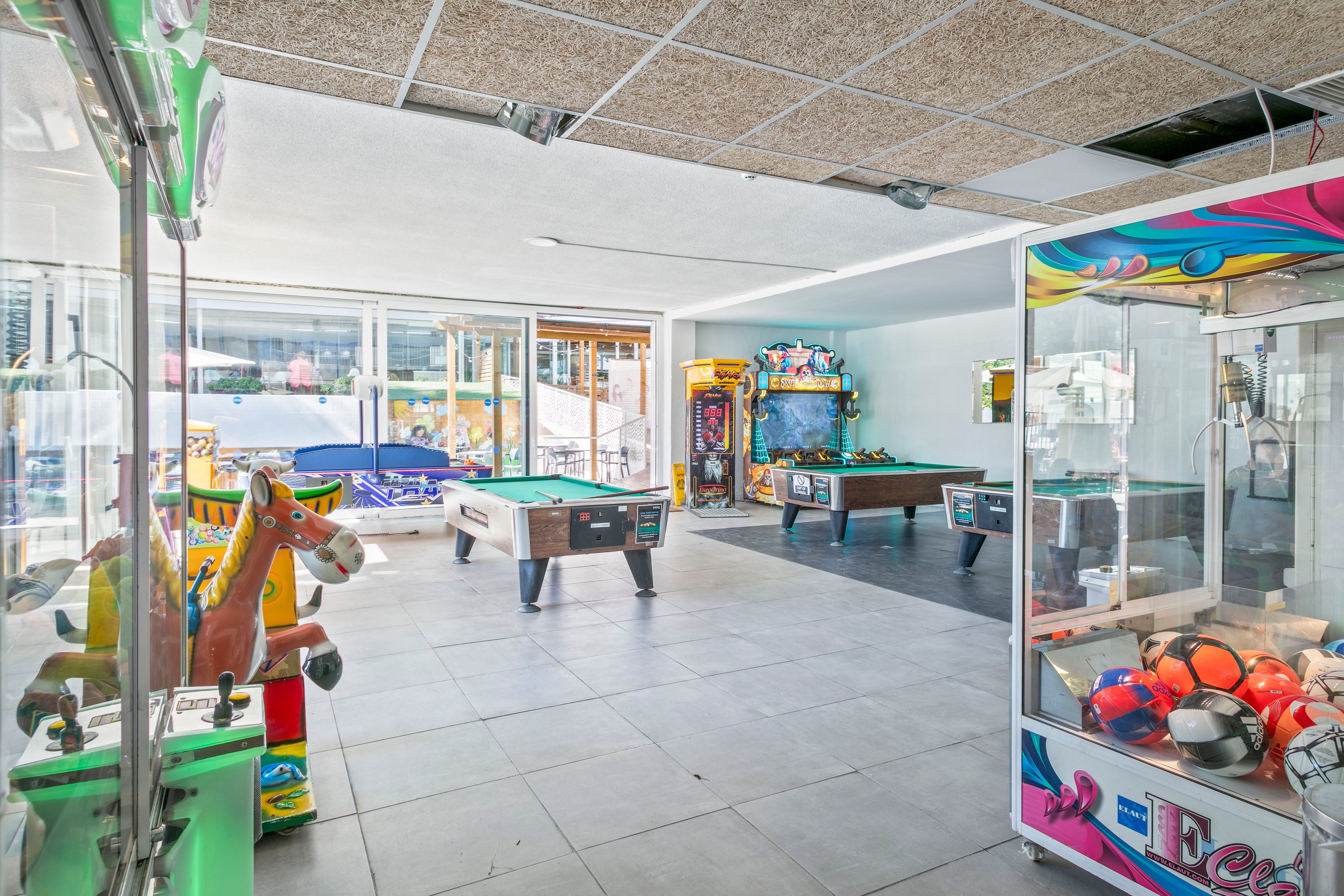 An indoor arcade room features pool tables, arcade machines, and claw cranes arranged on a tiled floor. Large windows along one wall let in natural light and reveal an outdoor area beyond. Colorful machines, including a kiddie ride shaped like a horse and a claw machine filled with balls, add a playful atmosphere. The space appears clean, modern, and ready for visitors to enjoy various games.