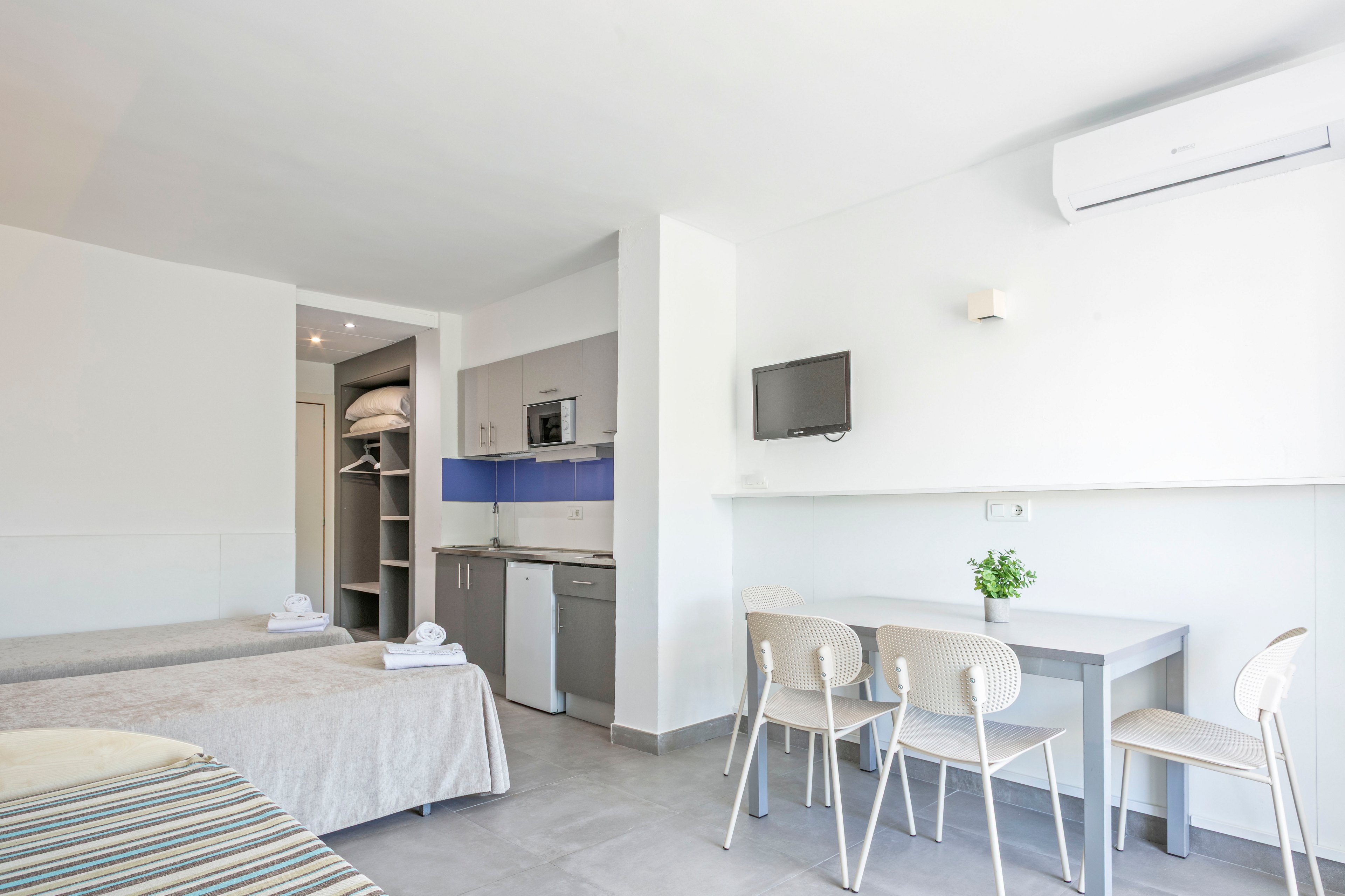 A bright studio apartment features two single beds neatly made with folded towels. The room includes a compact kitchenette with a small refrigerator, microwave, and open shelving. A wall-mounted television and air conditioning unit provide modern comforts above a simple dining table with four chairs. Neutral tones, clean lines, and a small green plant create a fresh, minimalist atmosphere.