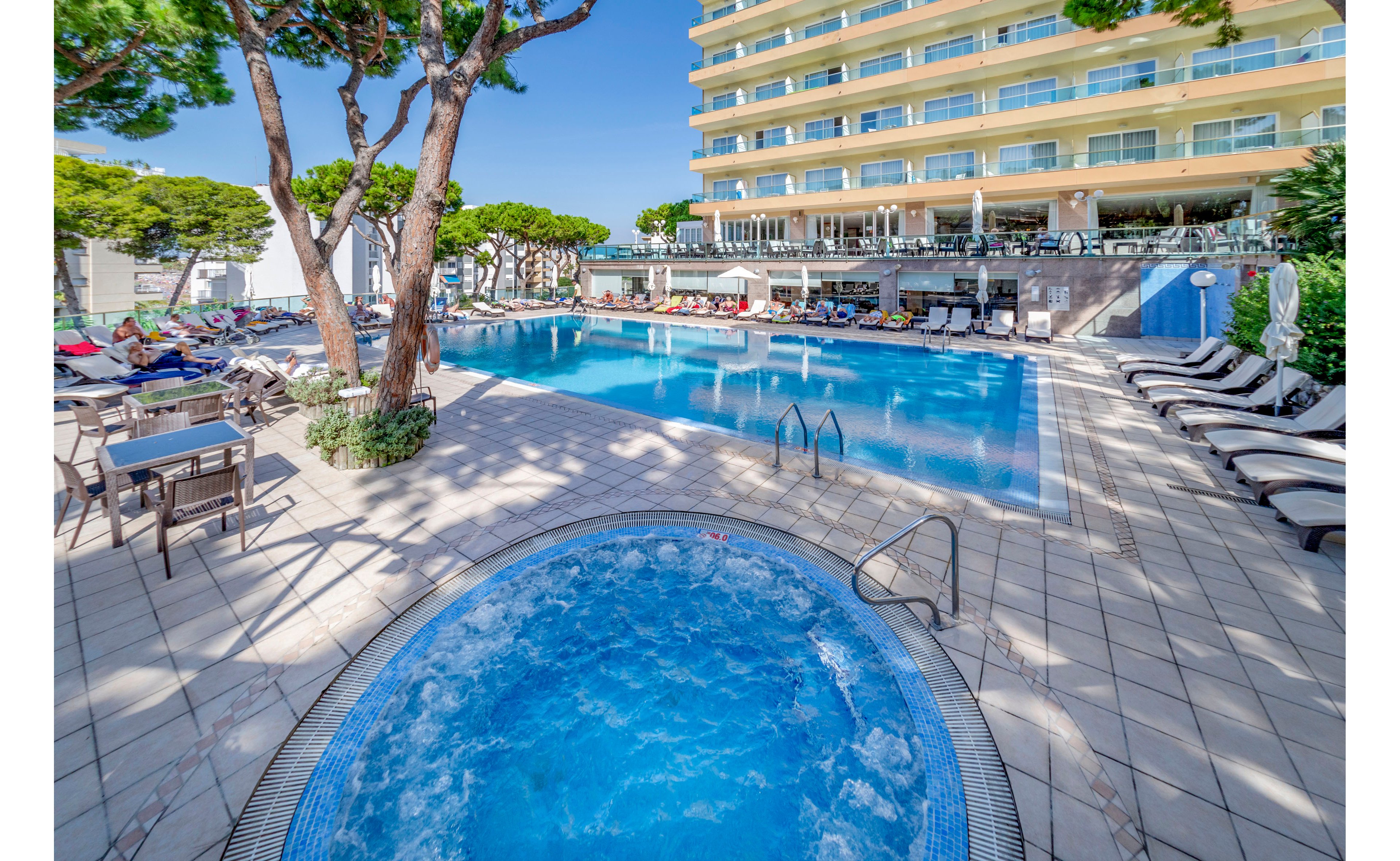 A modern hotel features a large outdoor swimming pool with a circular jacuzzi in the foreground. Sun loungers and umbrellas line the tiled pool deck, creating a relaxing resort atmosphere. A multi-story hotel building with balconies overlooks the pool area, while tall green trees add natural shade. Guests can be seen enjoying the water and lounging around the pool in bright daylight.