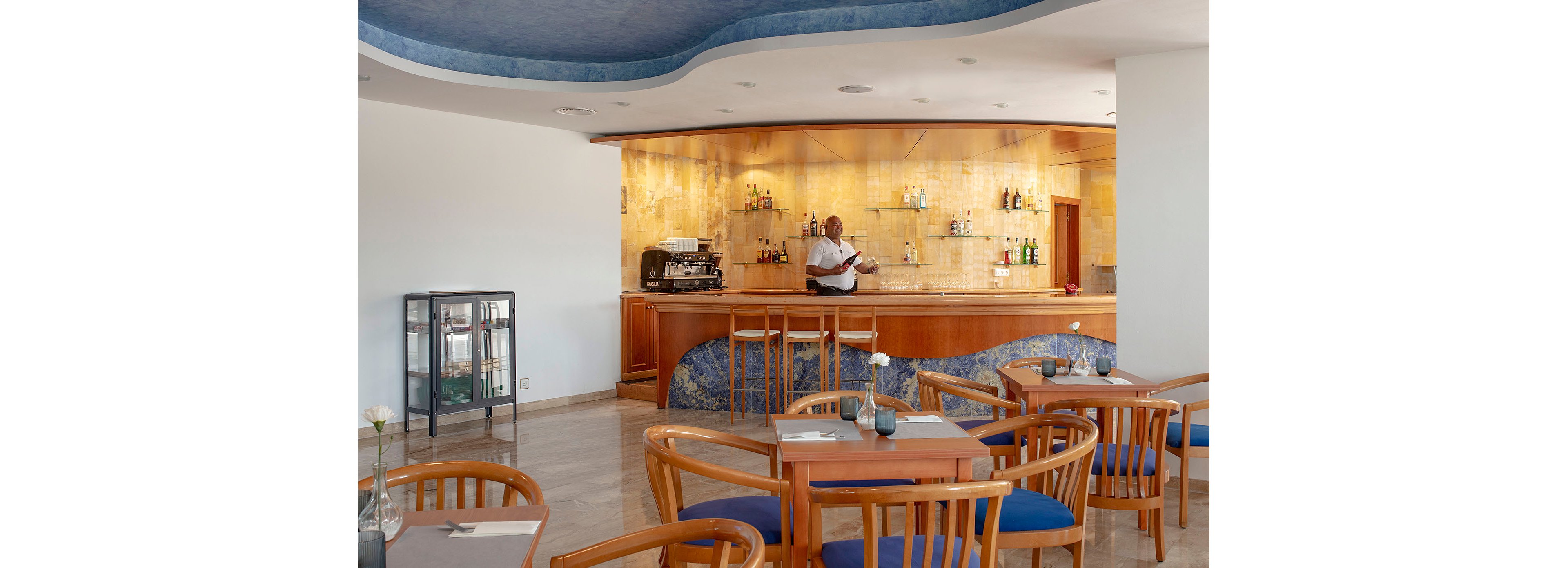 A bartender stands behind a polished wooden bar in a modern hotel lounge. The bar back wall is tiled in warm golden tones and displays various bottles and glassware. Wooden tables and chairs with blue cushions are arranged neatly in the foreground, each with simple table settings and small vases. The space is bright and clean, with a curved blue ceiling adding a contemporary touch.