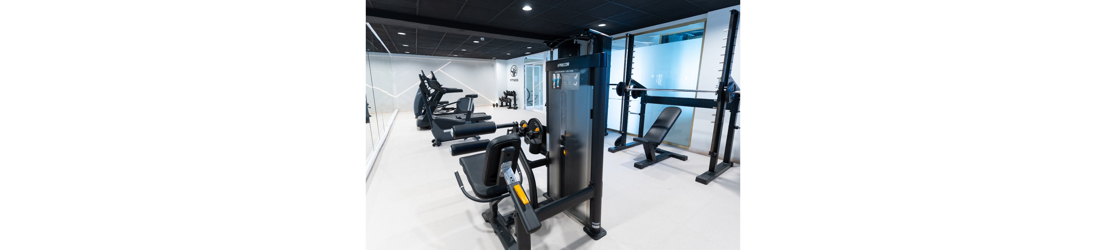 A clean, modern gym interior features several strength and cardio machines neatly arranged in a bright room. In the foreground, a seated leg exercise machine faces a large multi-function weight station. Treadmills and other cardio equipment line the mirrored wall on the left, while a bench press station sits to the right. The space is well-lit with a minimalist design, glass partitions, and light-colored flooring, creating an organized and professional fitness environment.