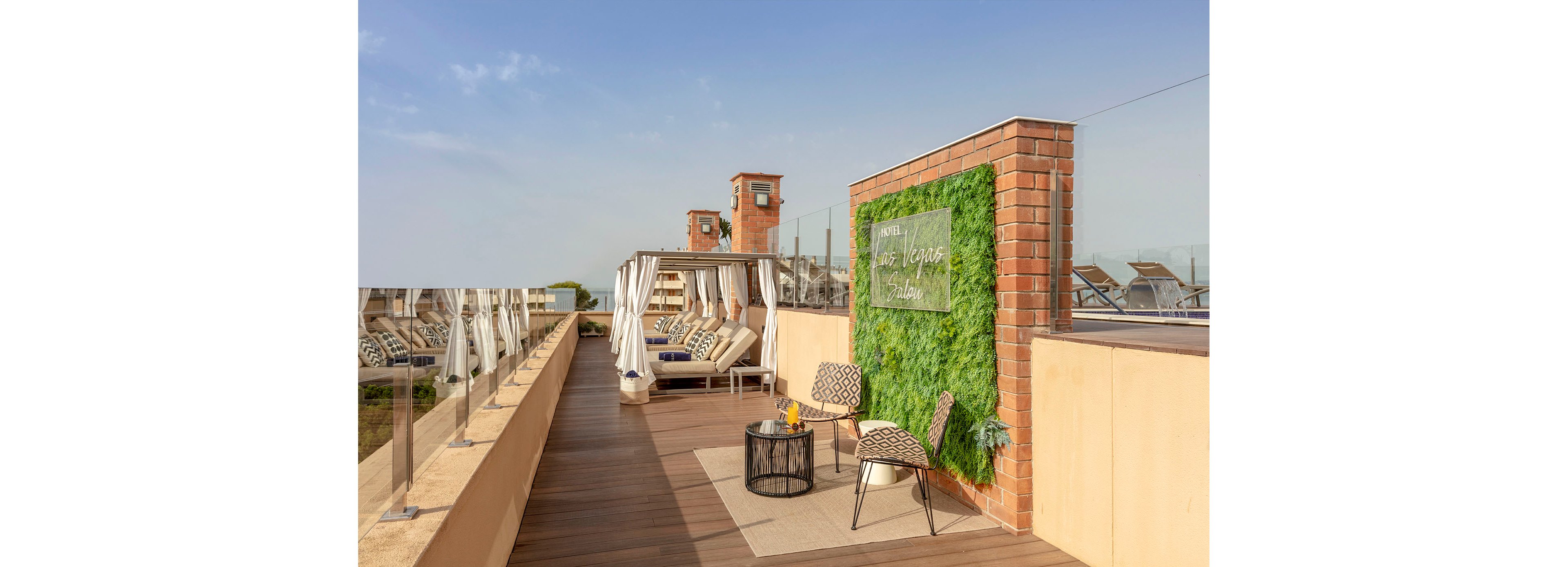 A stylish rooftop lounge features cabanas with cushioned seating arranged along a wooden deck. In the foreground, a small seating area includes patterned chairs and a round table with decorative items. A vertical green wall with a translucent sign adds a modern touch beside a brick column. Sun loungers line the far side of the terrace under a clear, bright sky, creating a relaxed outdoor retreat.