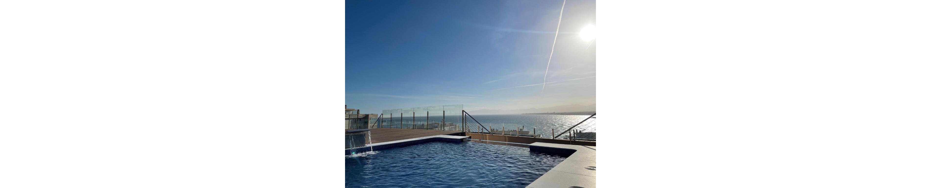 A modern rooftop pool sits beneath a clear blue sky, overlooking a calm ocean horizon. Glass railings and a wooden deck frame the pool area, giving an elevated, contemporary feel. Sunlight reflects off the water surface, creating shimmering highlights and a tranquil atmosphere. In the distance, faint landforms and buildings line the coast under the bright afternoon sun.