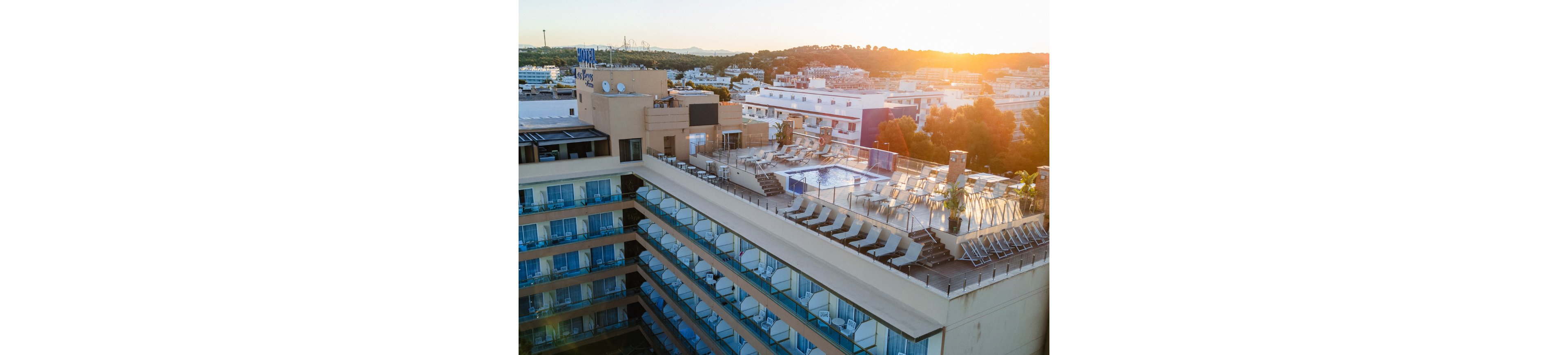 An elevated view shows a modern multi-story hotel with a rooftop swimming pool. Numerous lounge chairs surround the pool area, suggesting a relaxing leisure space. The building features rows of balconies with blue-tinted glass railings overlooking the city. In the background, low-rise buildings, trees, and distant hills are visible under a warm sunset sky.