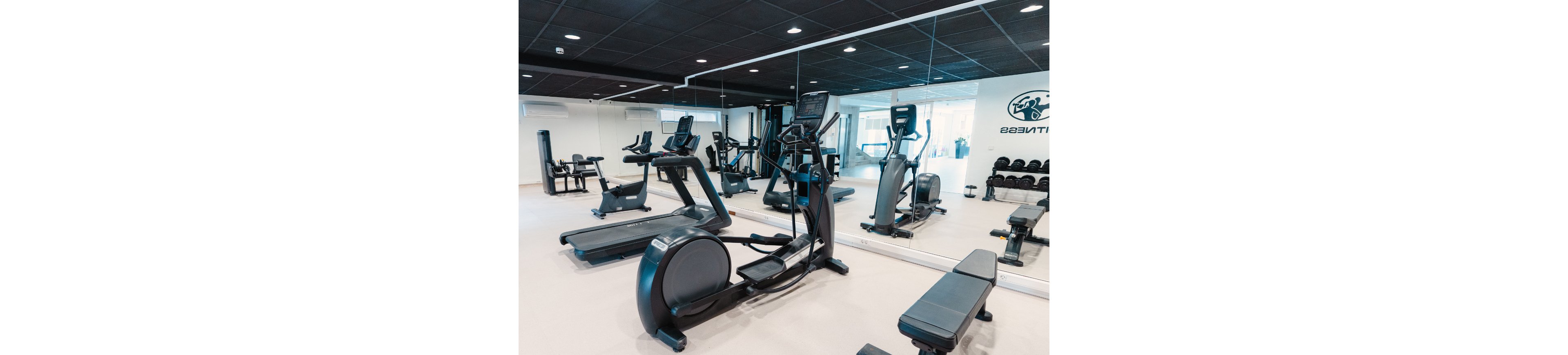A clean, modern gym features several treadmills and elliptical machines arranged in front of a large wall mirror. Weight benches and dumbbells are visible along the side, creating a full fitness area. The bright interior has light flooring, white walls, and a dark ceiling for contrast. No people are present, emphasizing the spacious and organized workout environment.