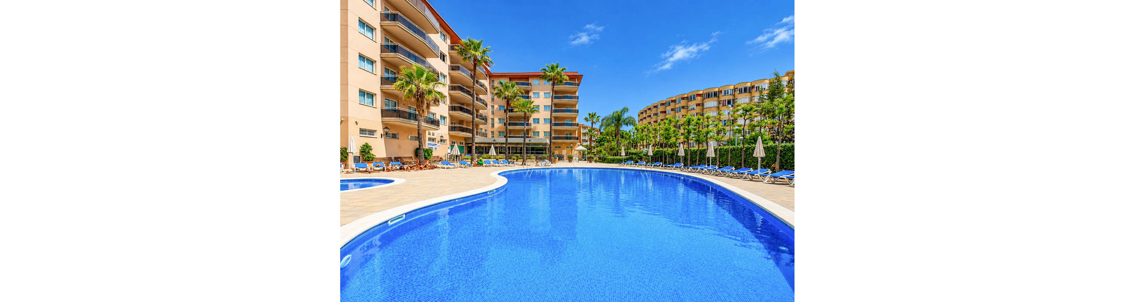 A large curved swimming pool dominates the foreground of a sunny resort scene. Multi-story apartment or hotel buildings surround the pool, with balconies overlooking the water. Sun loungers and umbrellas line the tiled pool deck, suggesting a relaxing vacation atmosphere. Tall palm trees and lush greenery add a tropical feel under a bright blue sky.