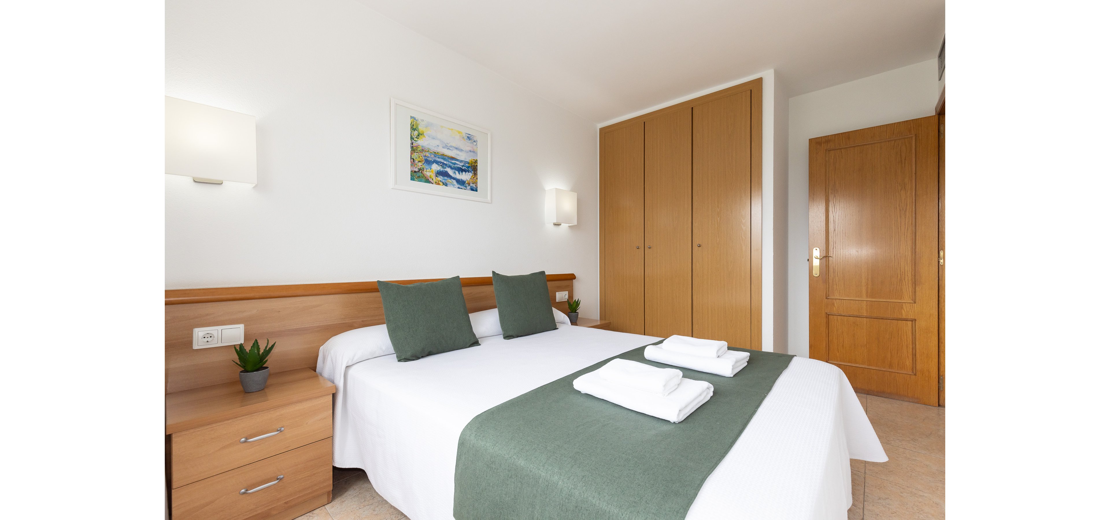 A bright, neatly arranged bedroom features a double bed with white linens and green accent pillows and runner. Folded white towels are placed on the bed, suggesting a hotel or guest accommodation. Wooden furniture, including bedside tables and a large built-in wardrobe, adds warmth to the clean, white walls. Soft wall lighting and a framed landscape painting contribute to the calm, inviting atmosphere.