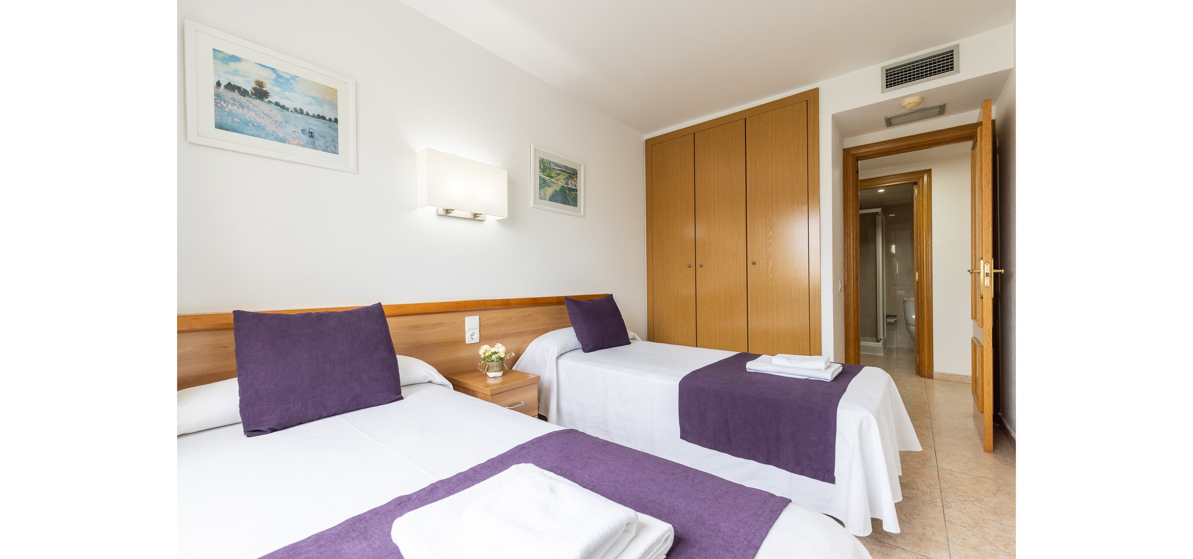 Neatly made twin beds with white linens and purple runners are arranged in a bright, modern hotel room. A wooden nightstand with a small plant sits between the beds, and folded towels are placed at the foot of each bed. Built-in wooden wardrobes line one wall, while an open door reveals a hallway leading to the bathroom. Soft wall lighting and framed artwork contribute to the clean, welcoming atmosphere.