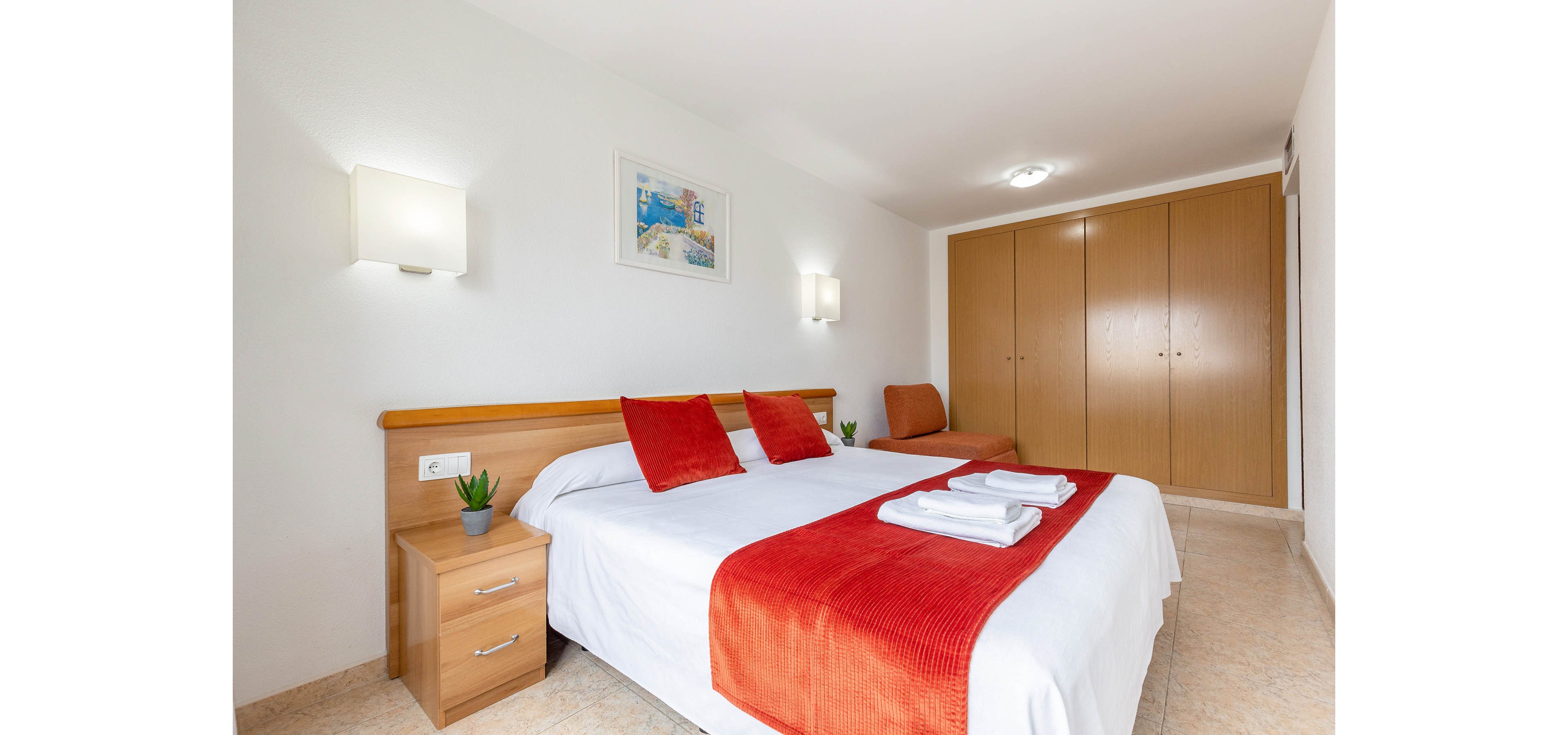 A clean, minimalist bedroom features a double bed with white linens and bold red throw blanket and pillows. The room is well lit with wall-mounted lamps and natural light, creating an airy atmosphere. Wooden furniture, including bedside tables, a large wardrobe, and a chair, adds warmth to the space. A framed artwork and small potted plants provide subtle decorative touches.