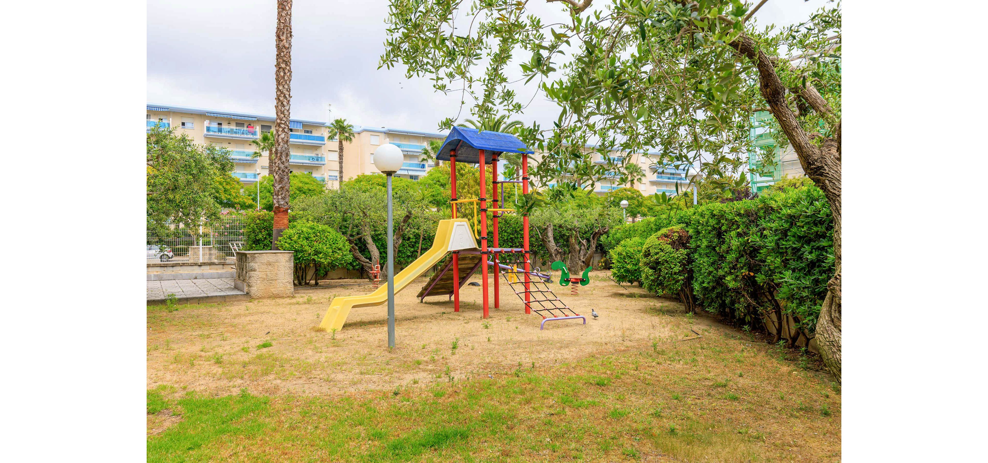 A small playground with a colorful climbing structure and slide stands in a grassy courtyard. The play equipment features a yellow slide, red and blue bars, and a small roofed platform. Surrounding the playground are trimmed bushes, trees, and a white globe streetlamp. In the background, multi-story residential buildings with balconies overlook the quiet outdoor space.