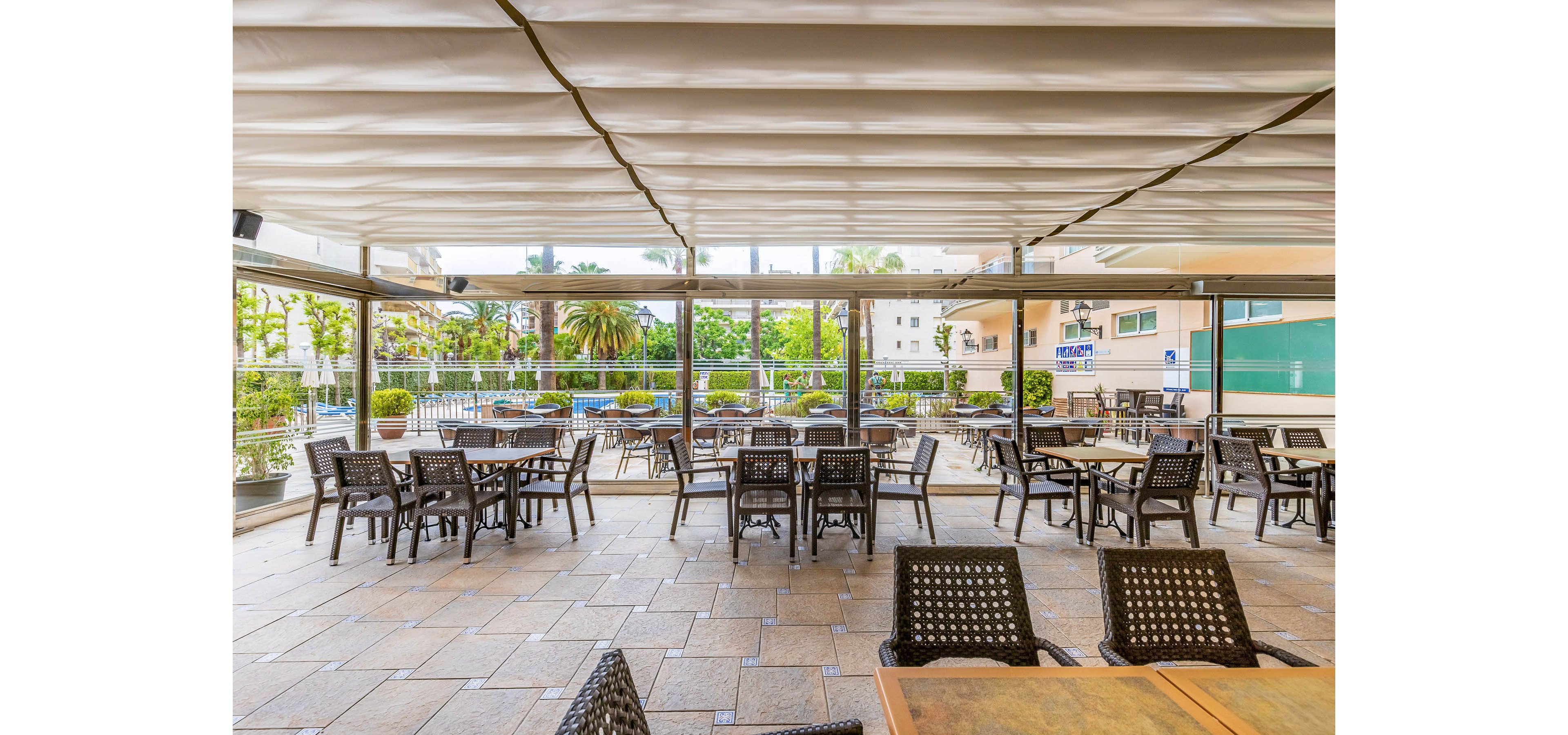 A spacious outdoor cafe terrace features multiple empty tables and dark woven chairs arranged on a tiled floor. The area is covered by a retractable canopy and enclosed with glass panels, offering views of a pool and lush greenery outside. In the background, palm trees, shrubs, and nearby buildings create a relaxed urban resort atmosphere. The scene is brightly lit, suggesting a calm daytime setting before guests arrive.