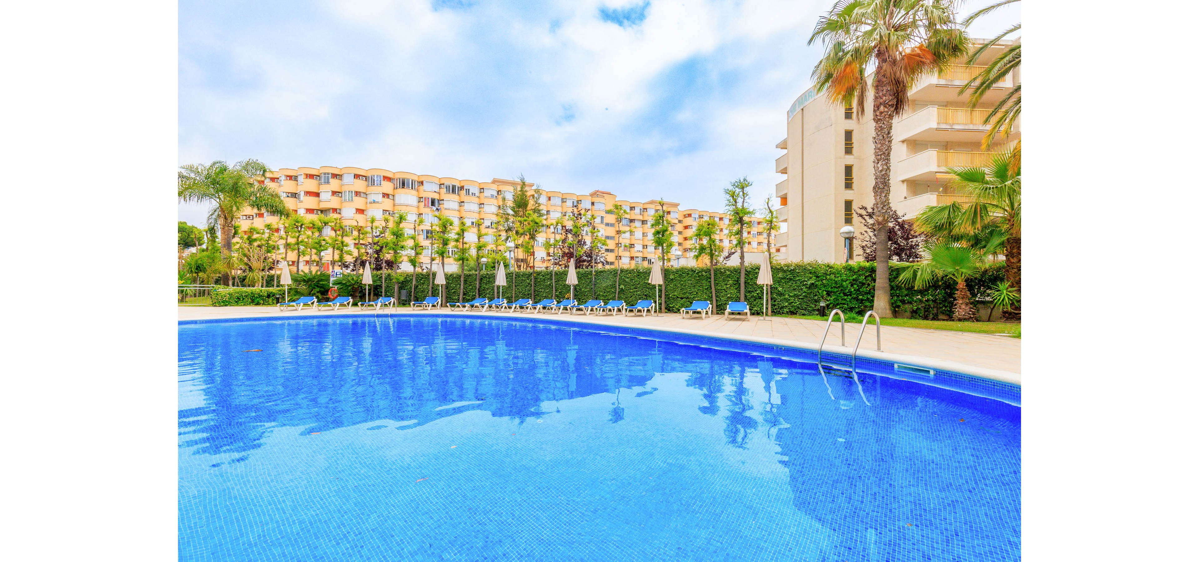 A large outdoor swimming pool dominates the foreground with bright blue water. Sun loungers and closed umbrellas line the poolside, suggesting a relaxing resort environment. In the background, multi-story apartment or hotel buildings rise behind neatly trimmed hedges and palm trees. The scene is captured on a partly cloudy day, creating a calm vacation atmosphere.