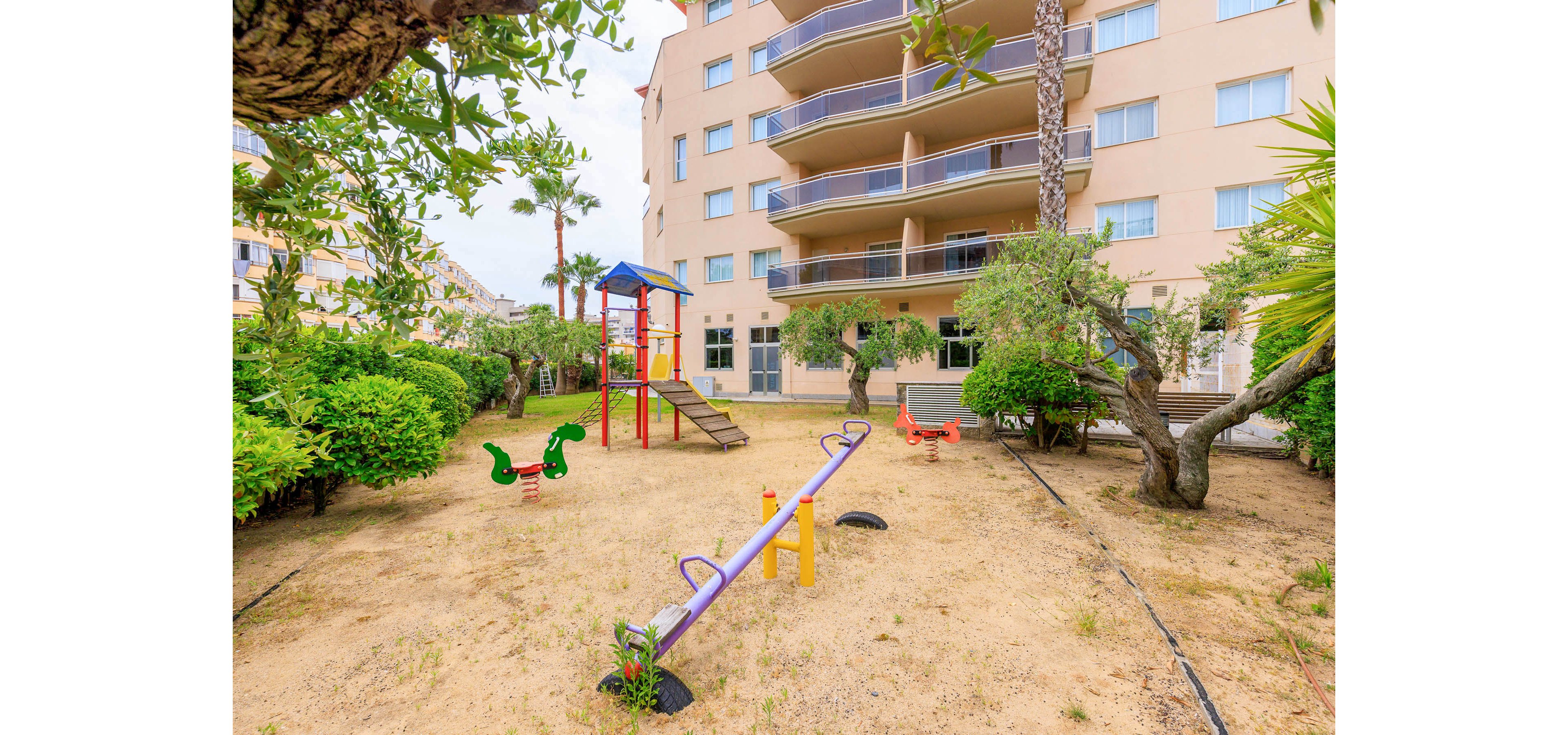 An outdoor playground is set in the courtyard of a multi-story residential building. The play area features a central seesaw, a slide tower with a ladder, and spring riders shaped like animals. Sandy ground contrasts with the surrounding green bushes, trees, and palm trees. Balconies and windows of the apartment building frame the background, suggesting a family-friendly urban environment.