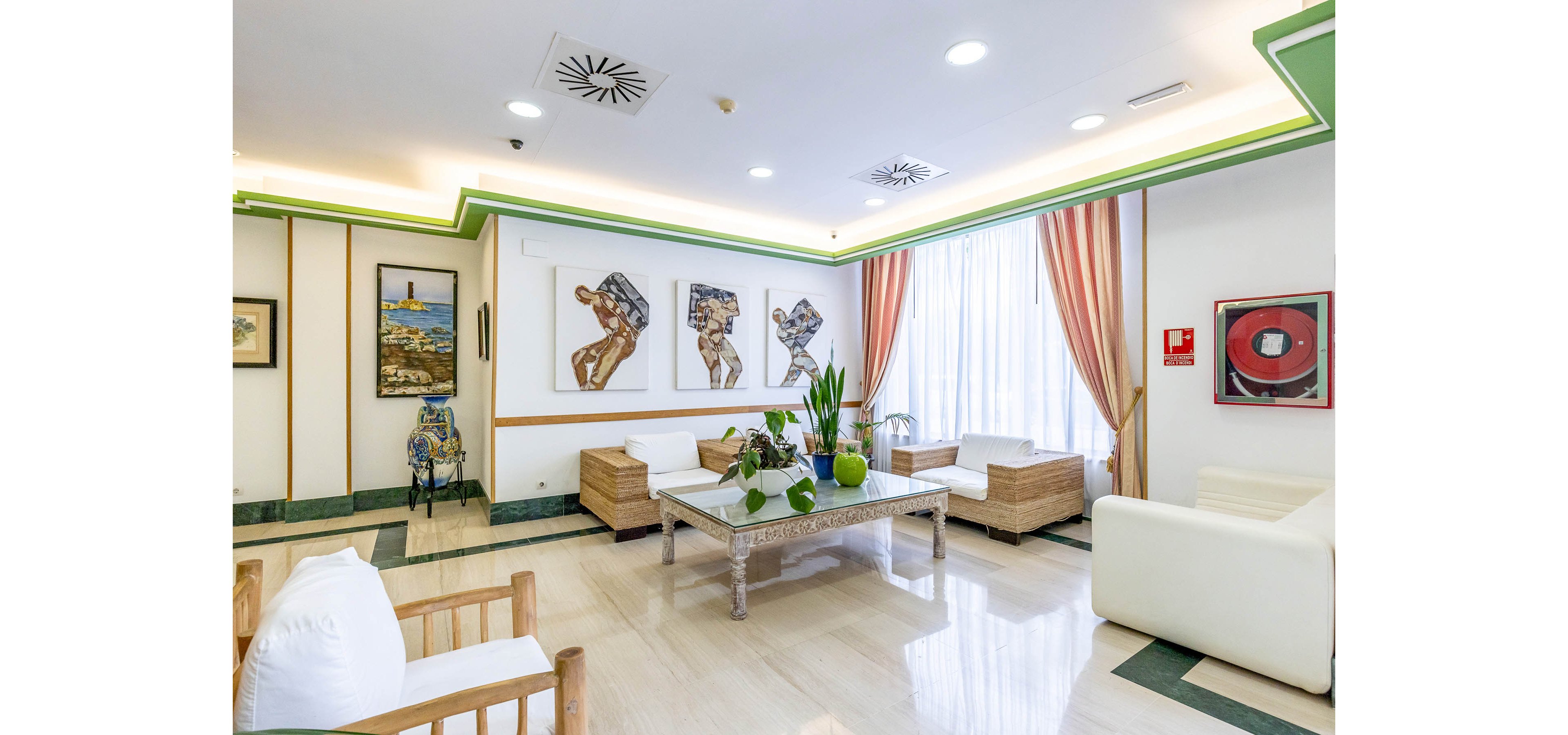 Spacious modern lounge area with white sofas and wicker armchairs is arranged around a decorative coffee table with green plants. Large windows with sheer curtains and peach drapes fill the room with natural light. Contemporary artwork and framed paintings decorate the walls, adding visual interest to the clean, minimalist design. The polished floor and recessed ceiling lighting contribute to the bright, welcoming atmosphere.