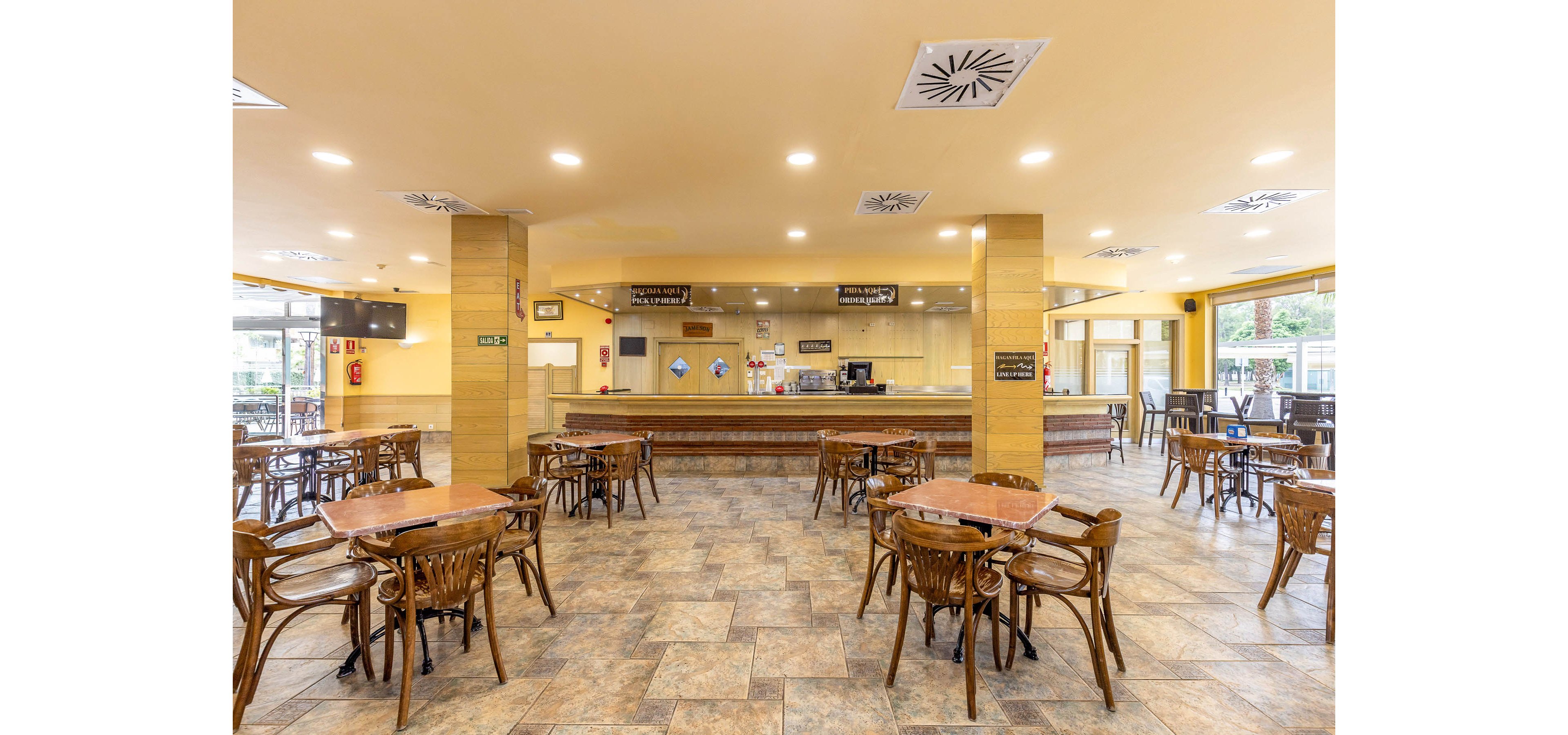 Spacious cafeteria interior with multiple wooden tables and chairs arranged across a tiled floor. A long service counter with coffee machines and display areas stretches along the back wall. Overhead menu boards and ceiling lights create a bright, welcoming atmosphere. Large windows on the right side let in natural light and offer views of greenery outside.
