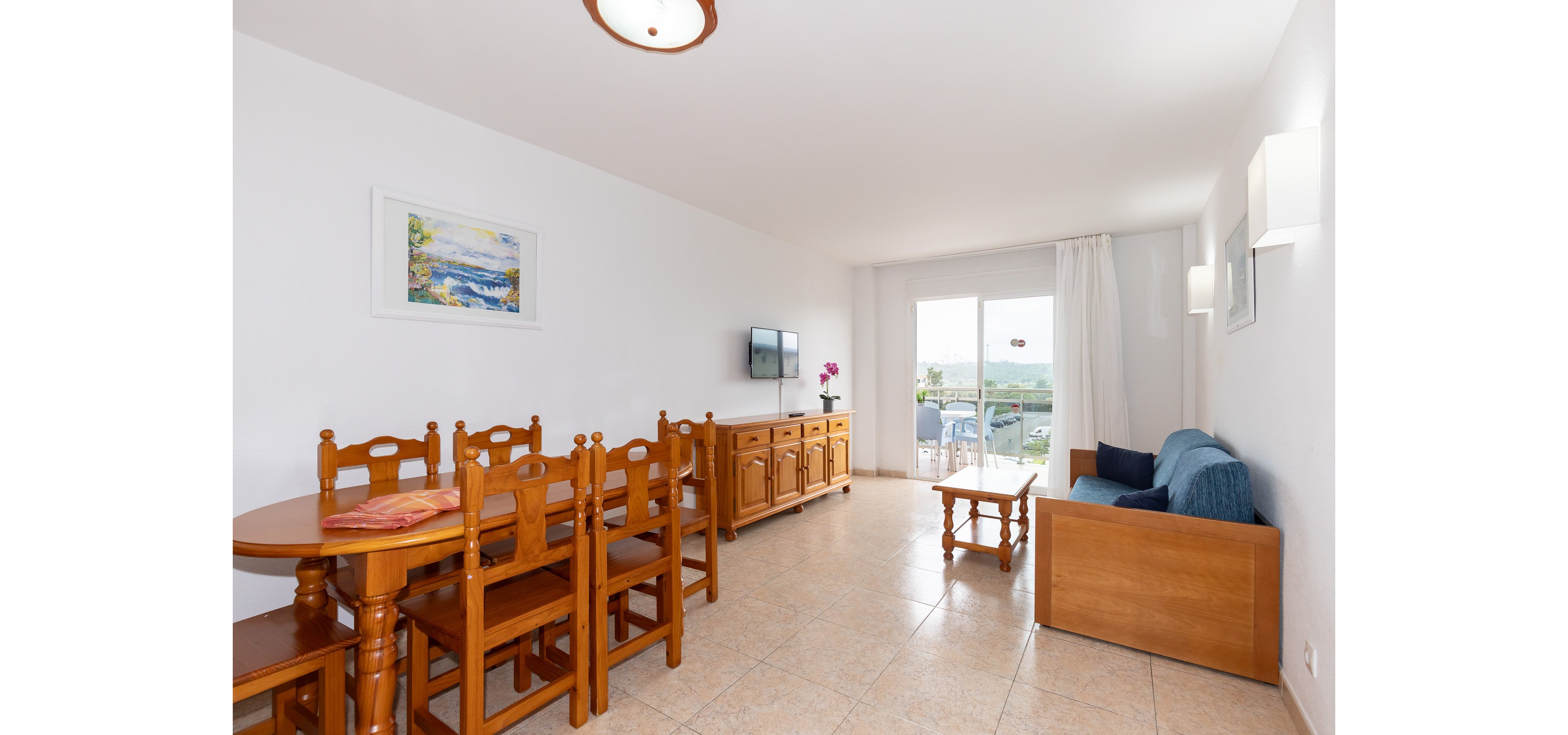 Spacious living and dining area in a bright holiday apartment with wooden furniture. A long dining table with several chairs stands beside a sideboard and wall-mounted television. A blue sofa and small coffee table face large sliding glass doors that open to a balcony with outdoor seating. Neutral tiled floors and white walls enhance the airy, minimalist feel of the room.