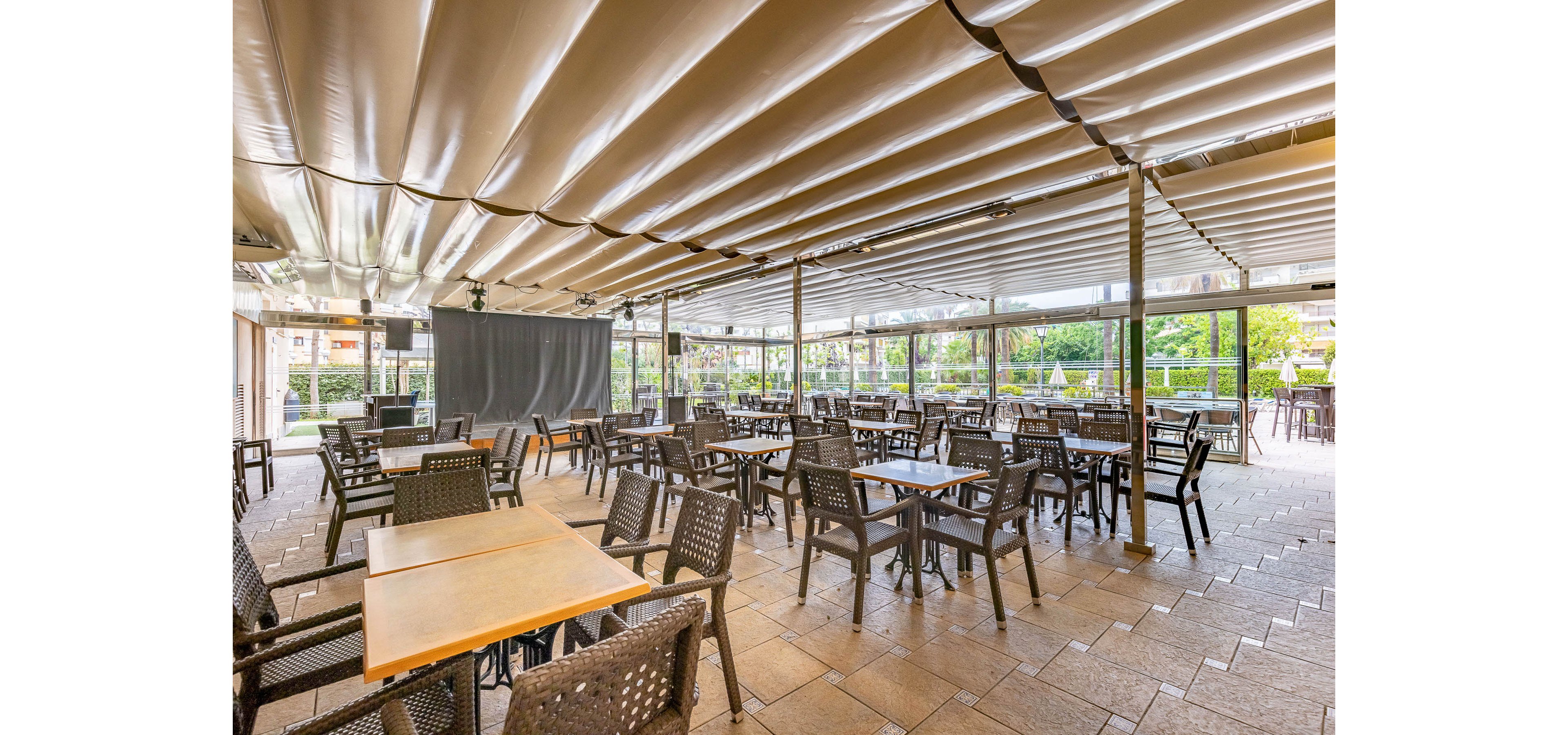 Spacious outdoor restaurant seating area with numerous empty tables and chairs is shown under a large retractable canopy. The setting appears to be a covered terrace or patio with tiled flooring and metal support columns. Natural light filters in from the open sides, revealing greenery and buildings in the background. The scene feels calm and ready for guests, suitable for dining or events.