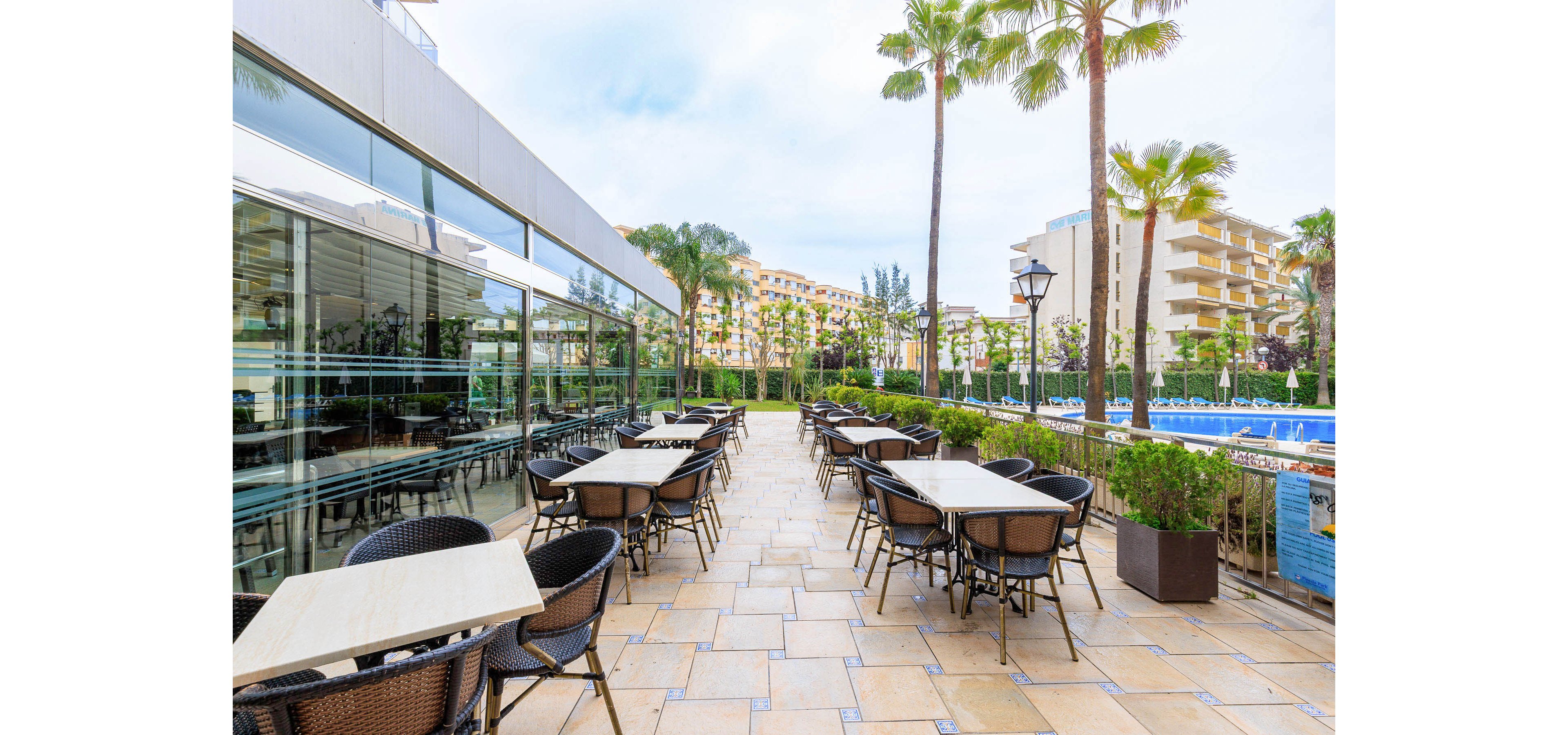 Spacious outdoor seating is arranged on a tiled hotel terrace beside a glass-walled building. Empty tables and wicker chairs line the walkway, creating a relaxed cafe or restaurant setting. A bright blue swimming pool and sun loungers are visible to the right, framed by tall palm trees. Modern apartment-style buildings rise in the background under a lightly overcast sky, suggesting a warm resort environment.