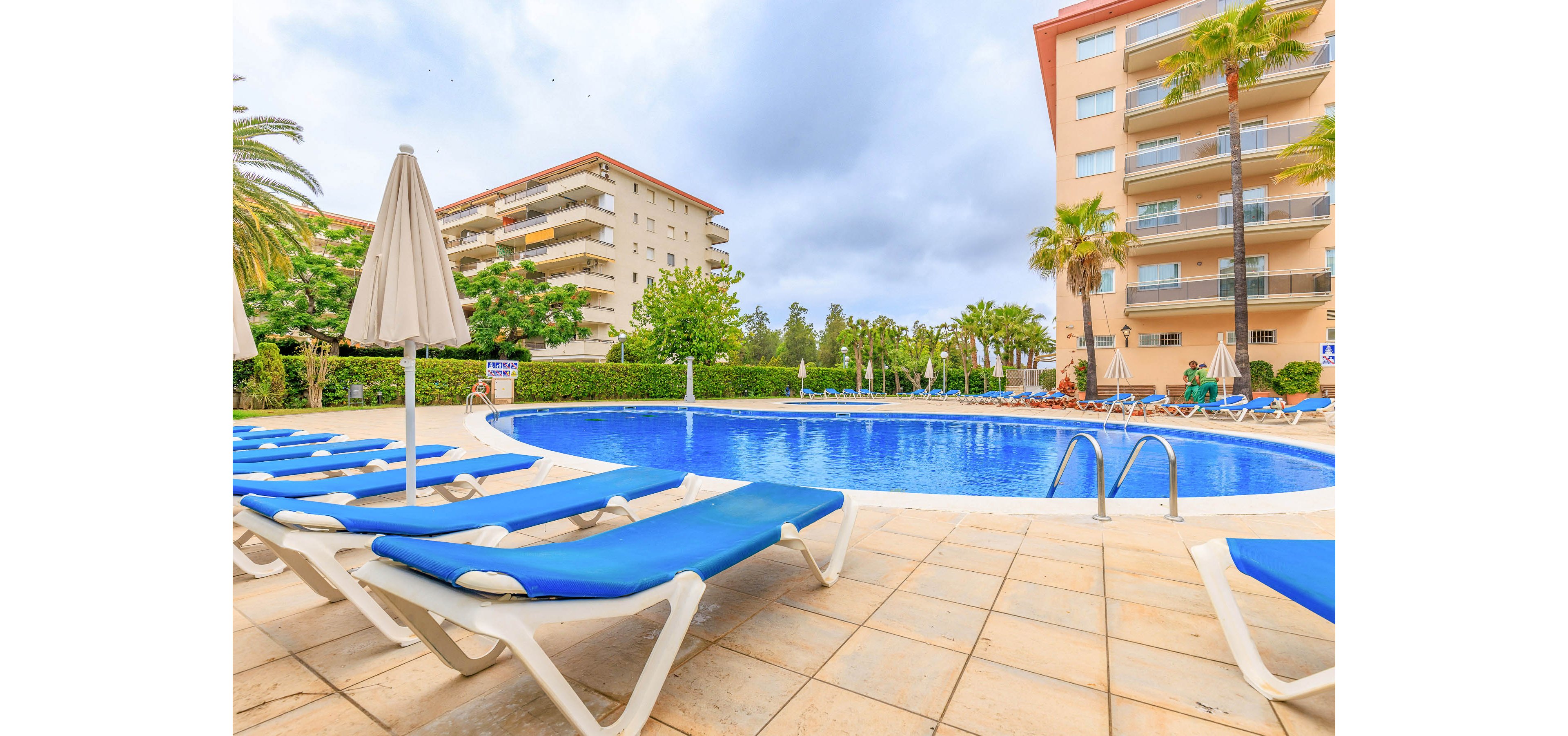 An outdoor resort swimming pool is surrounded by bright blue sun loungers and closed umbrellas. Apartment-style hotel buildings rise in the background under a cloudy sky. Palm trees and green hedges frame the pool area, creating a relaxed vacation atmosphere. The scene appears calm and empty, suggesting an early morning or off-peak moment at the resort.