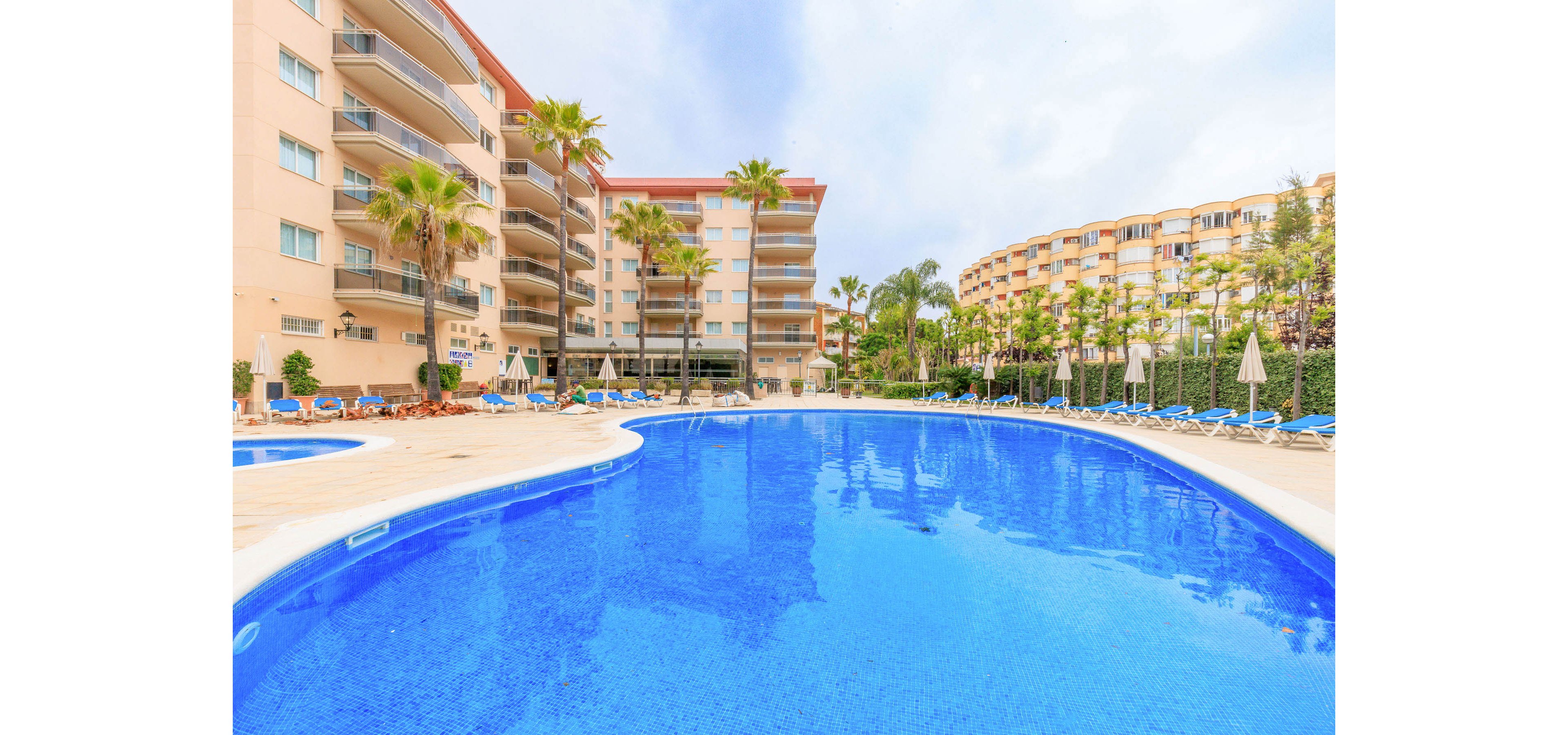 A large curved swimming pool dominates the foreground of a modern resort complex. Multi-story apartment buildings with balconies surround the pool area, lined with palm trees and sun loungers. Blue lounge chairs and white umbrellas are arranged along the pool deck, suggesting a relaxing vacation setting. The sky is lightly overcast, creating soft, even lighting across the scene.