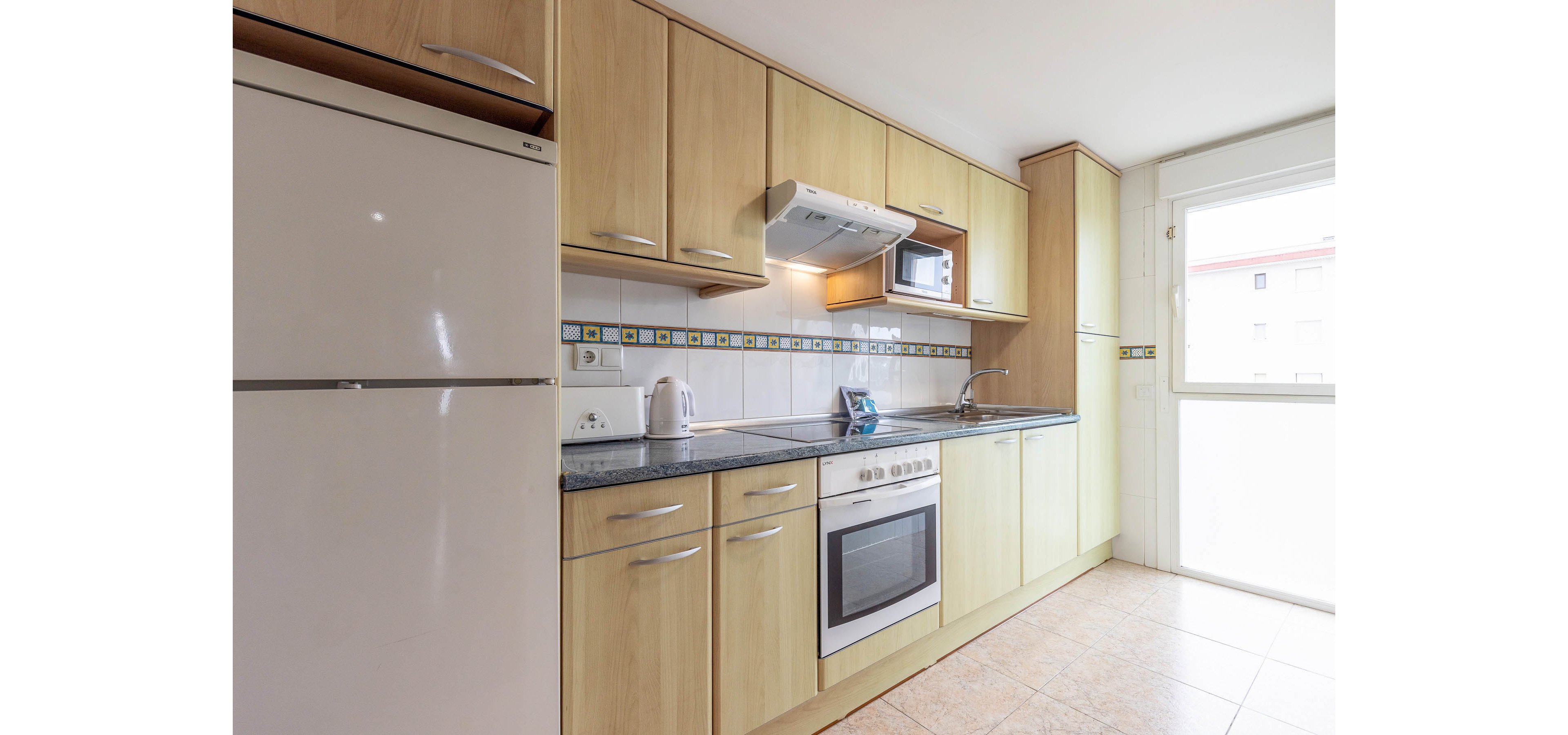 A bright modern kitchen features light wood cabinets and a dark countertop. Built-in appliances include an oven, stovetop, range hood, and a microwave. A white refrigerator stands to the left, with a kettle and toaster on the counter. Natural light enters through a large glass door, illuminating the tiled floor and backsplash with decorative tiles.