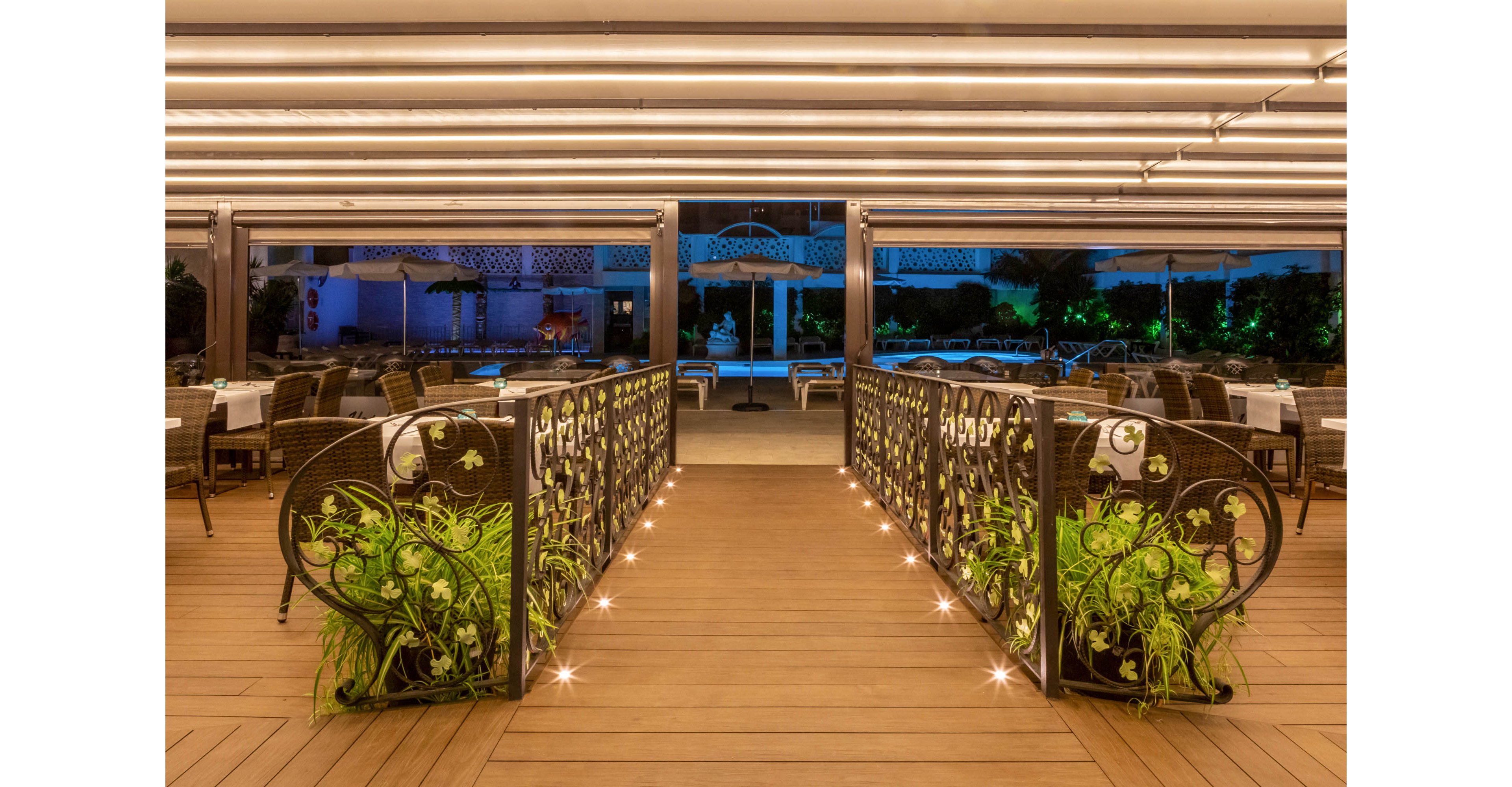 An upscale outdoor restaurant terrace features a decorative walkway leading toward a poolside area. Ornate railings with lush green plants line the wooden bridge, illuminated by small floor lights. Rows of dining tables and chairs are arranged neatly on both sides under a modern, striped ceiling. The scene is captured at night, with warm interior lighting contrasting against cool blue tones in the background.