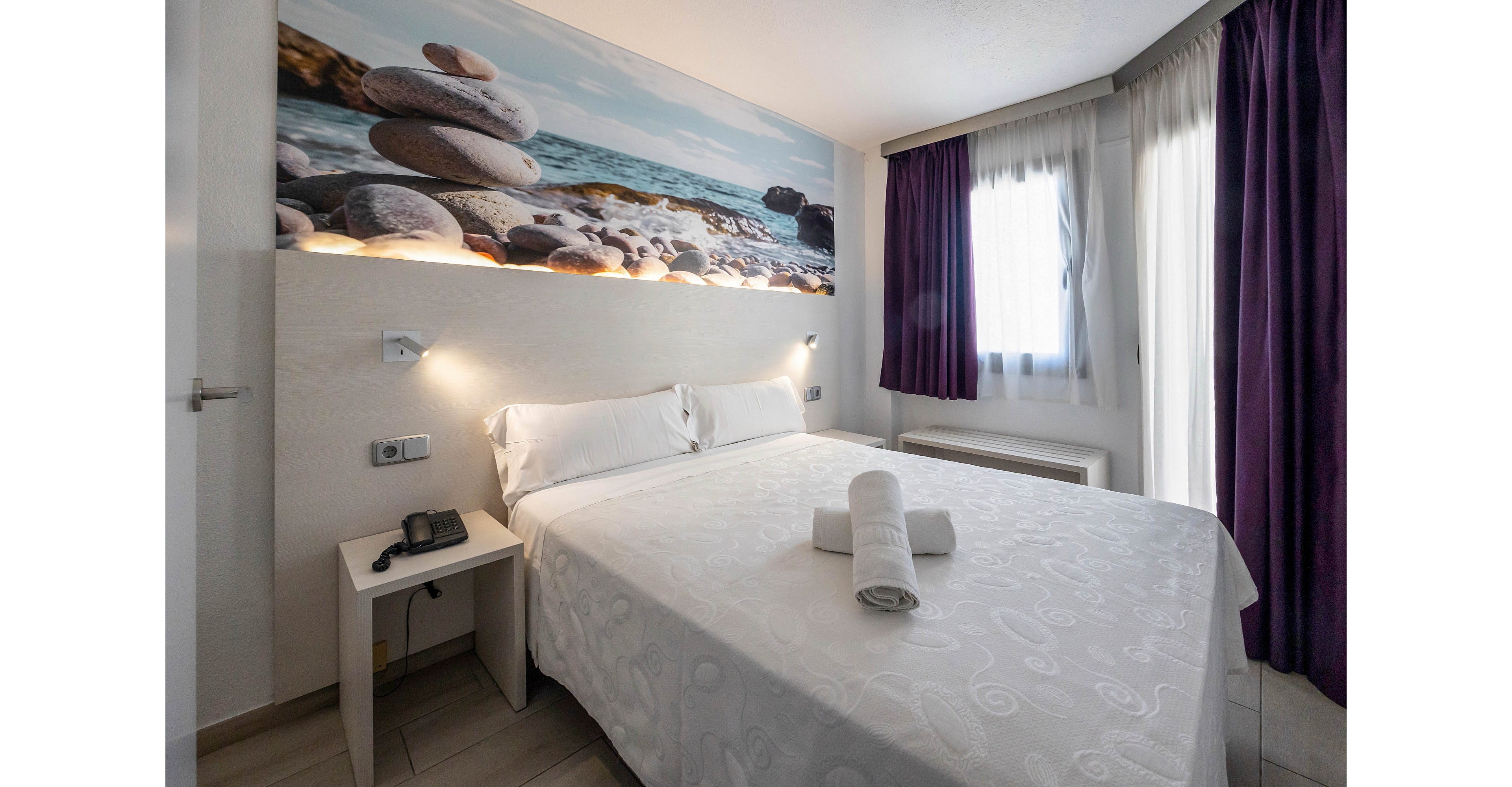 A neatly made double bed with white linens stands in a bright, minimalist hotel room. Rolled white towels are placed on the bed, adding a welcoming touch. Large windows with sheer curtains and dark drapes let in natural light, illuminating the space. A bedside table with a telephone and a large wall mural of stacked stones on a beach complete the serene, contemporary decor.
